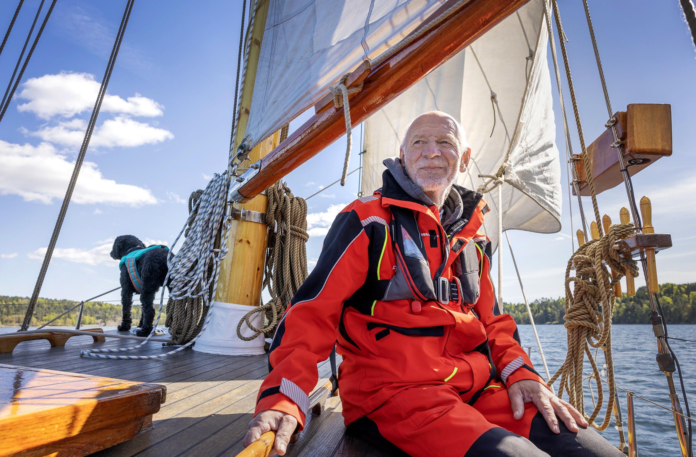 Foto: Mann i 60-årene uten hår og grått skjegg sitter på båten mens den seiler. I baugen står en svart hund og kikker utover vannet.