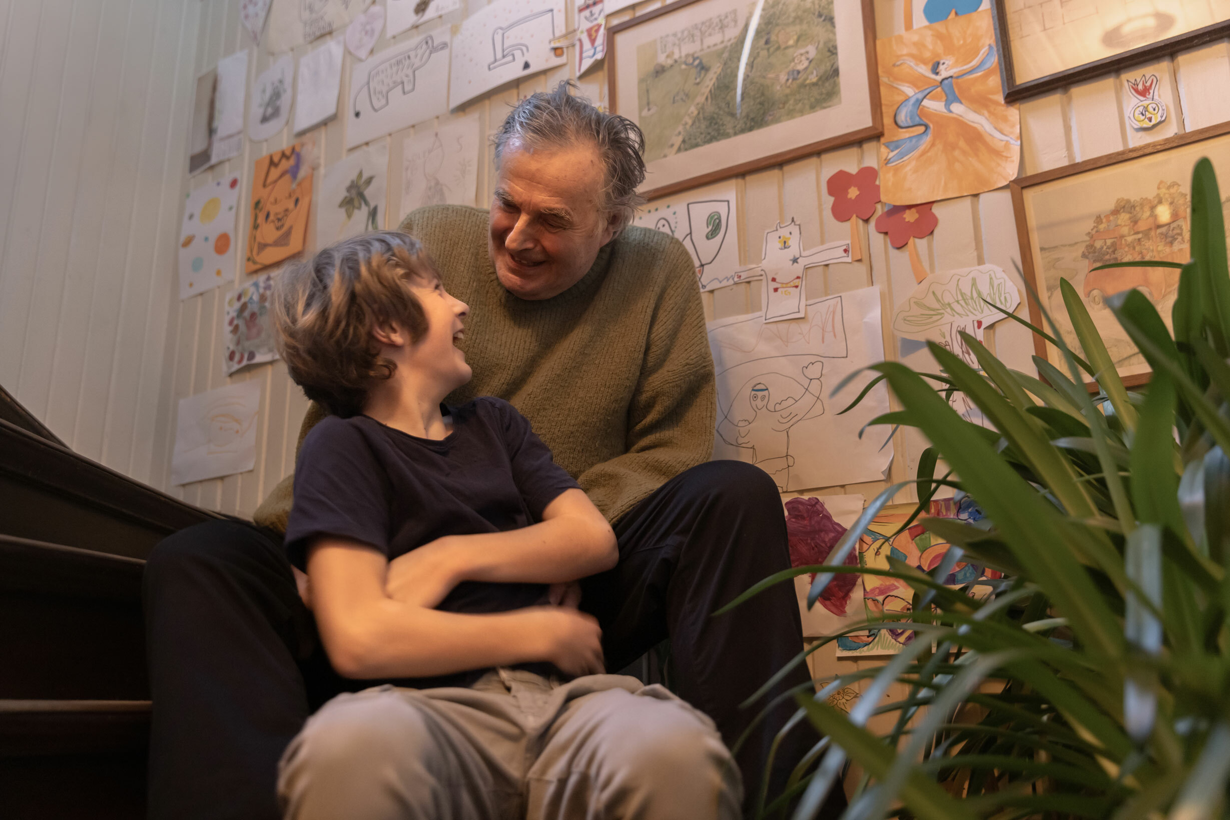 Bestefar og gutt på rundt 10 år sitter i en trapp. De ser p å hverandre og smiler. På veggen er det mange barnetegninger.