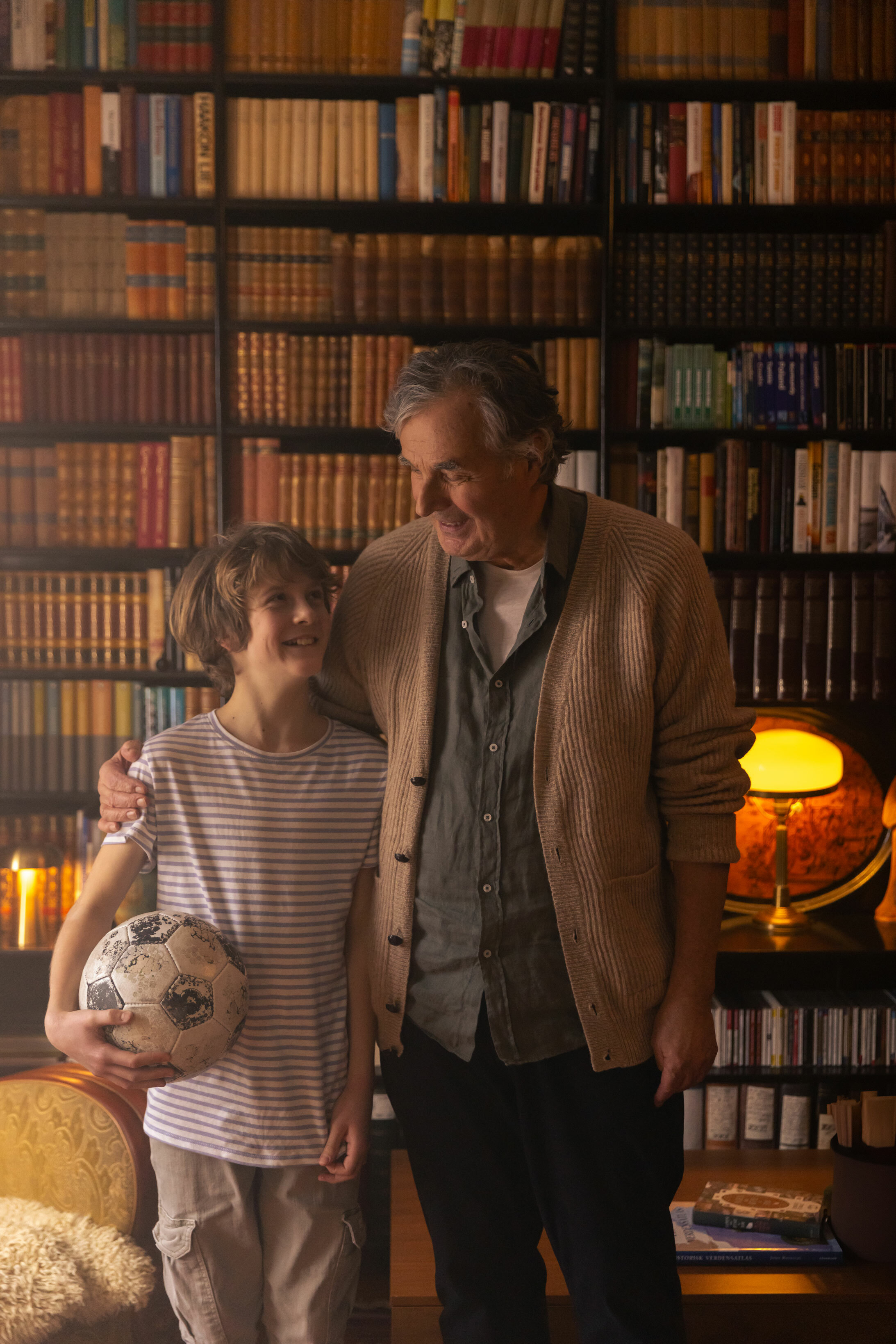 Bestefar og barnebarn står i et bibliotek sammen. Gutten holder en fotball.