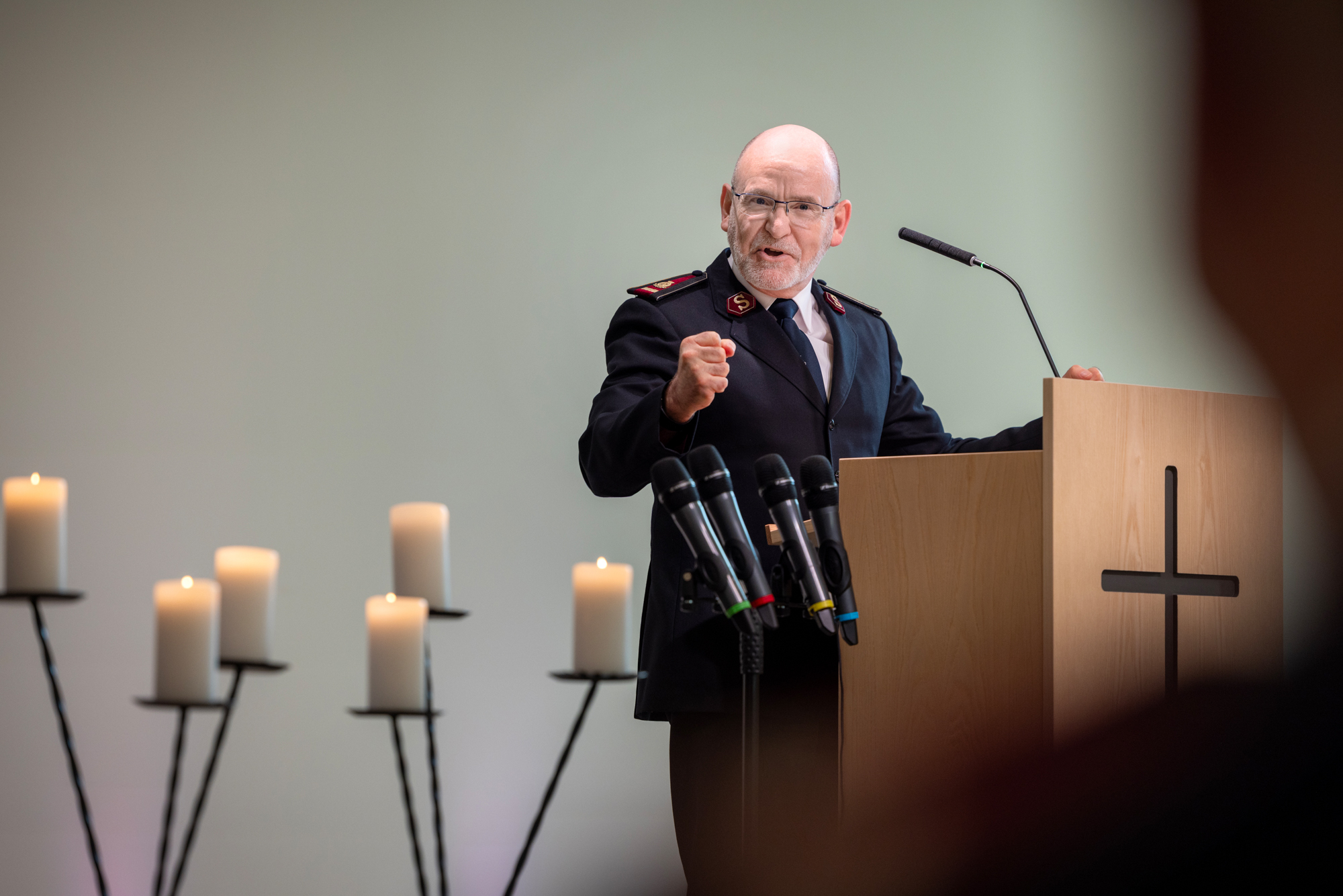 Foto: Mann på talerstolen med knyttede hender. Han bruker briller og er uten hår. Står på en talerstol og har på seg uniform fra Frelsesarmeen.