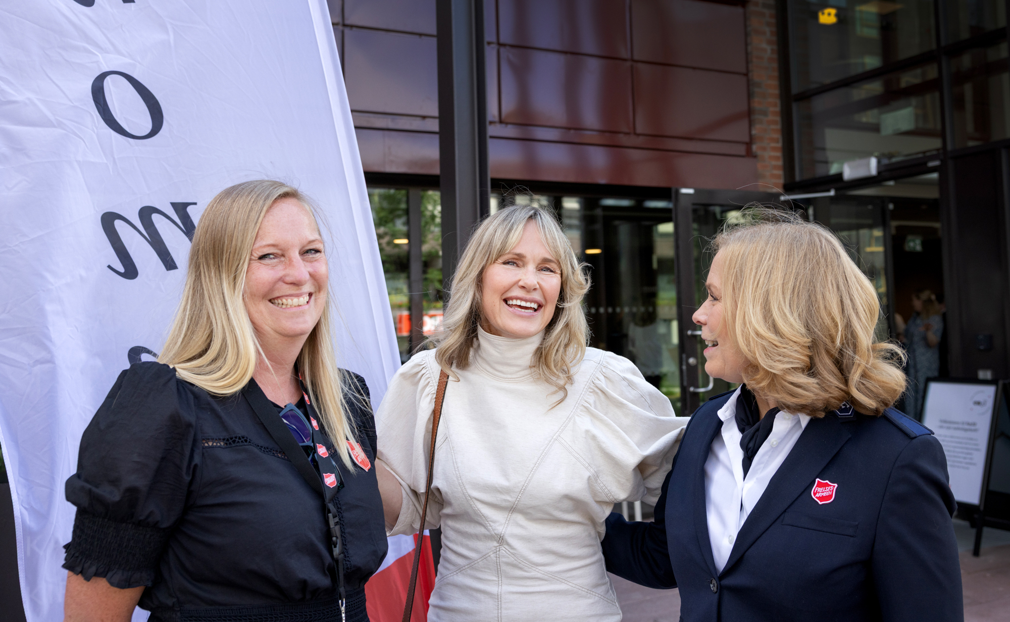 Foto: Tre blonde kvinner i 50-årene står tett sammen utenfor et kontorbygg med en "flagg" der det står velkommen.