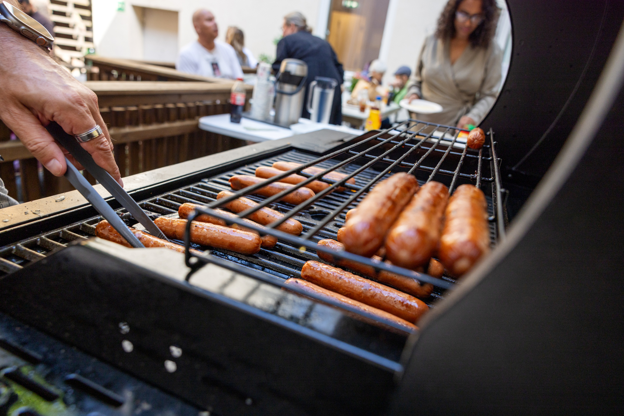 Et nærbilde av en grill med mange grillpølser på. Vi ser så vidt en hånd med egnet utstyr som snur på pølsene.