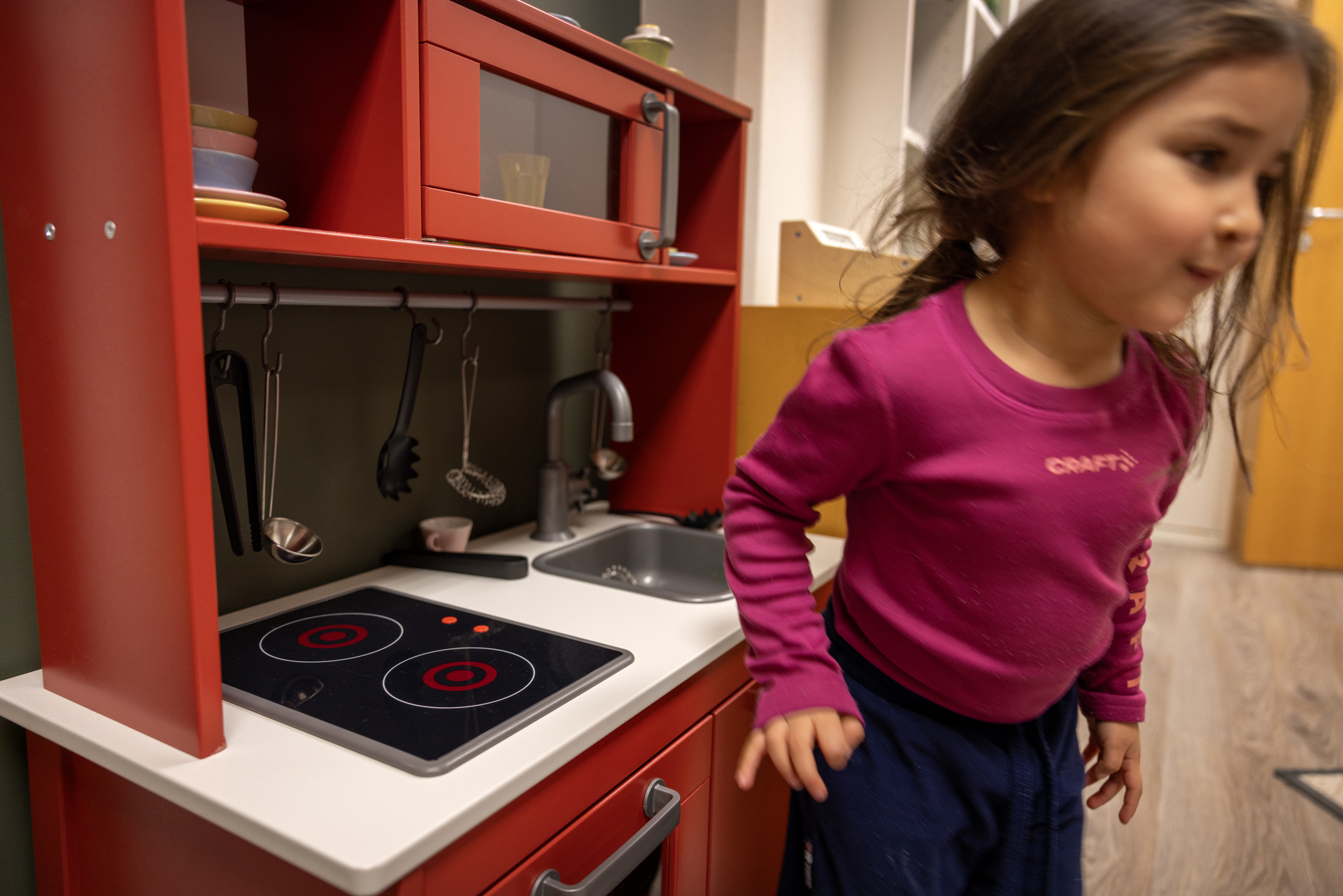 Foto: To bilder etter hverandre der det første viser en liten jente som hopper ned fra en klatrevegg med fargerike grep å holde i. Den andre viser samme jente som står ved en rød lekekomfyr.