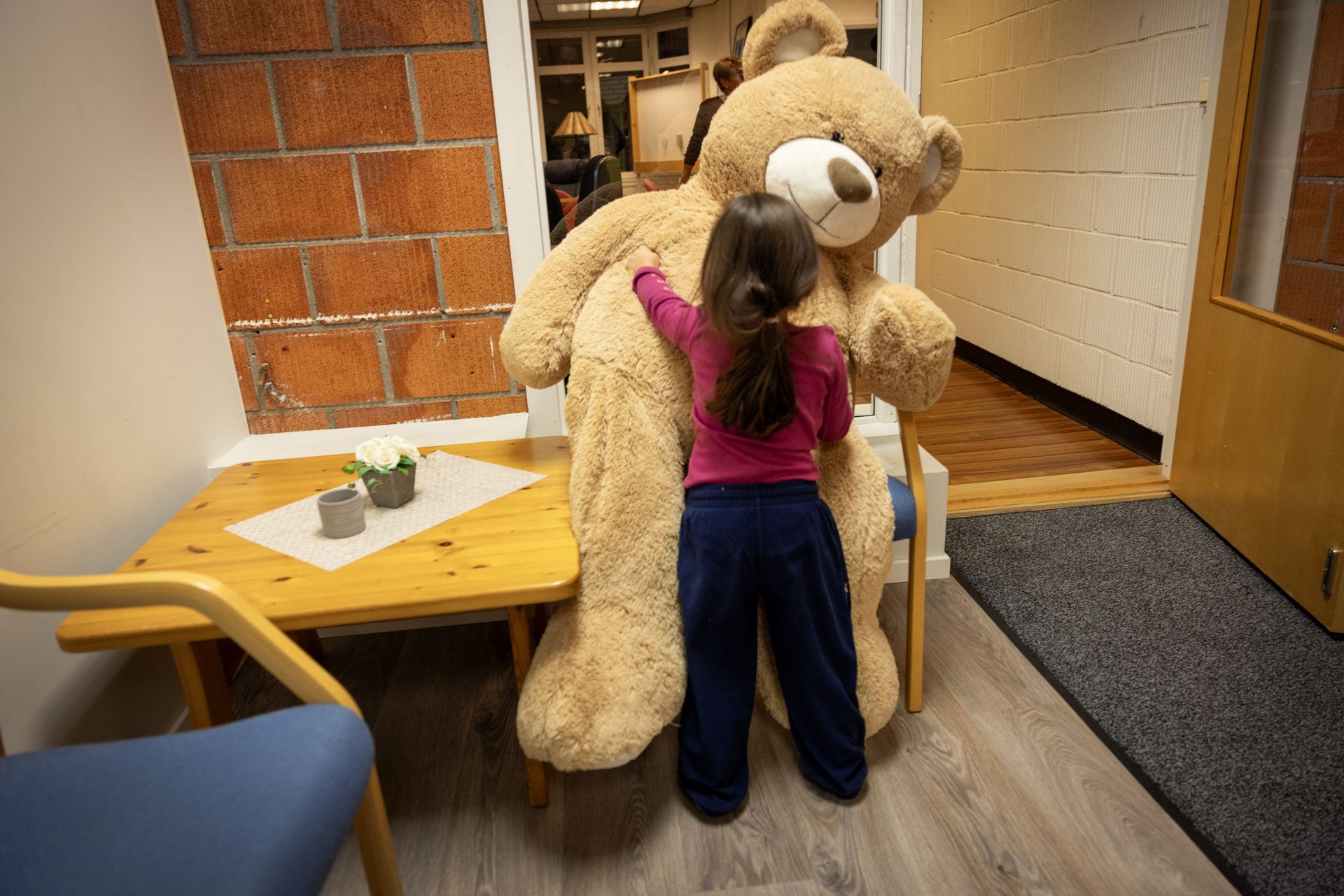 Foto: Liten jente med mørkt, langt hår som står med ryggen til og koser en stor bamse.