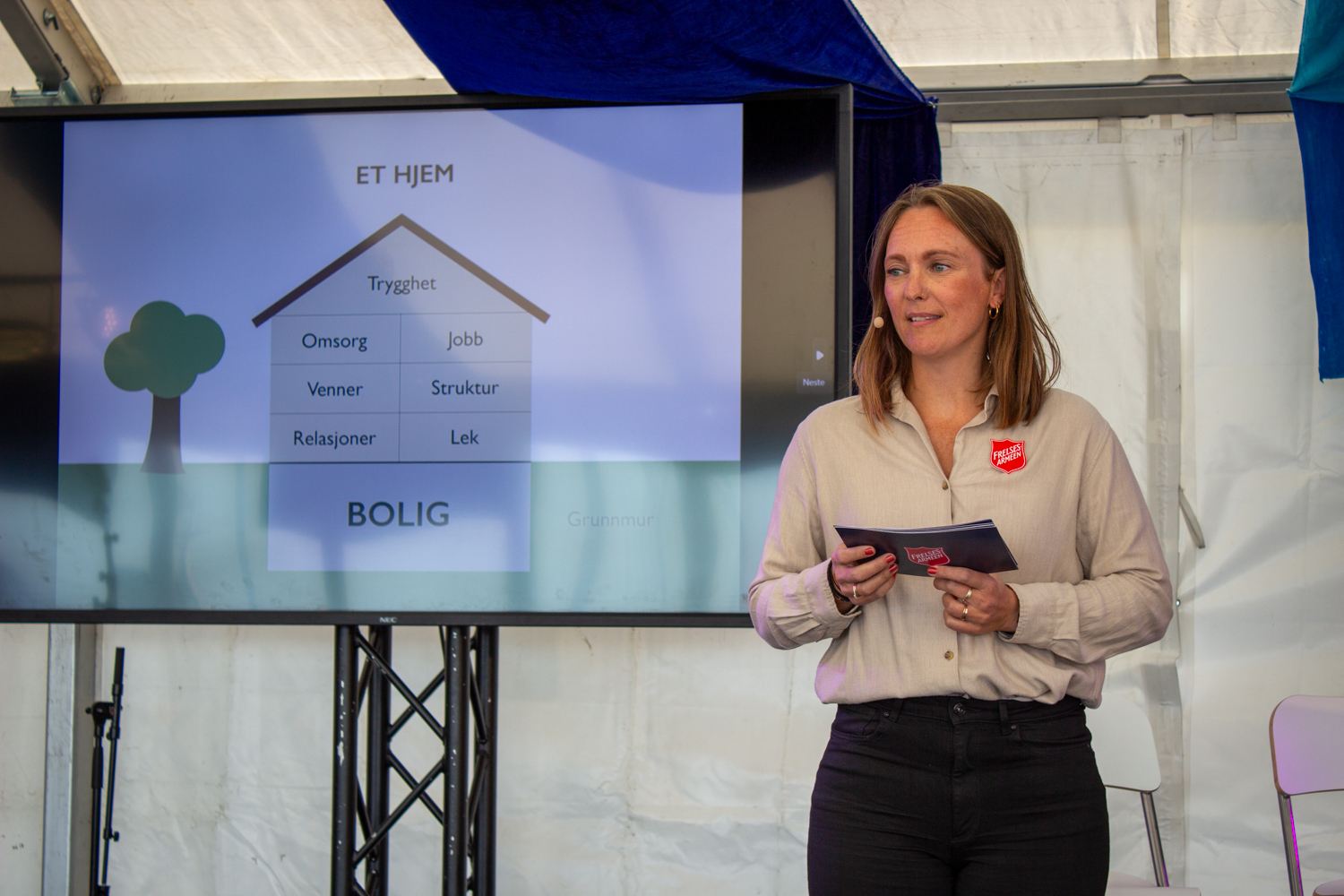 Foto: Kvinne med halvlangt, mørkeblondt hår står i beige skjorte og svart bukse på en scene. Den er i et stort utendørs telt for bak ser vi teltduken i hvite og blå striper. På skjermen viser hun et hus der det står tekst om hva et hjem er.