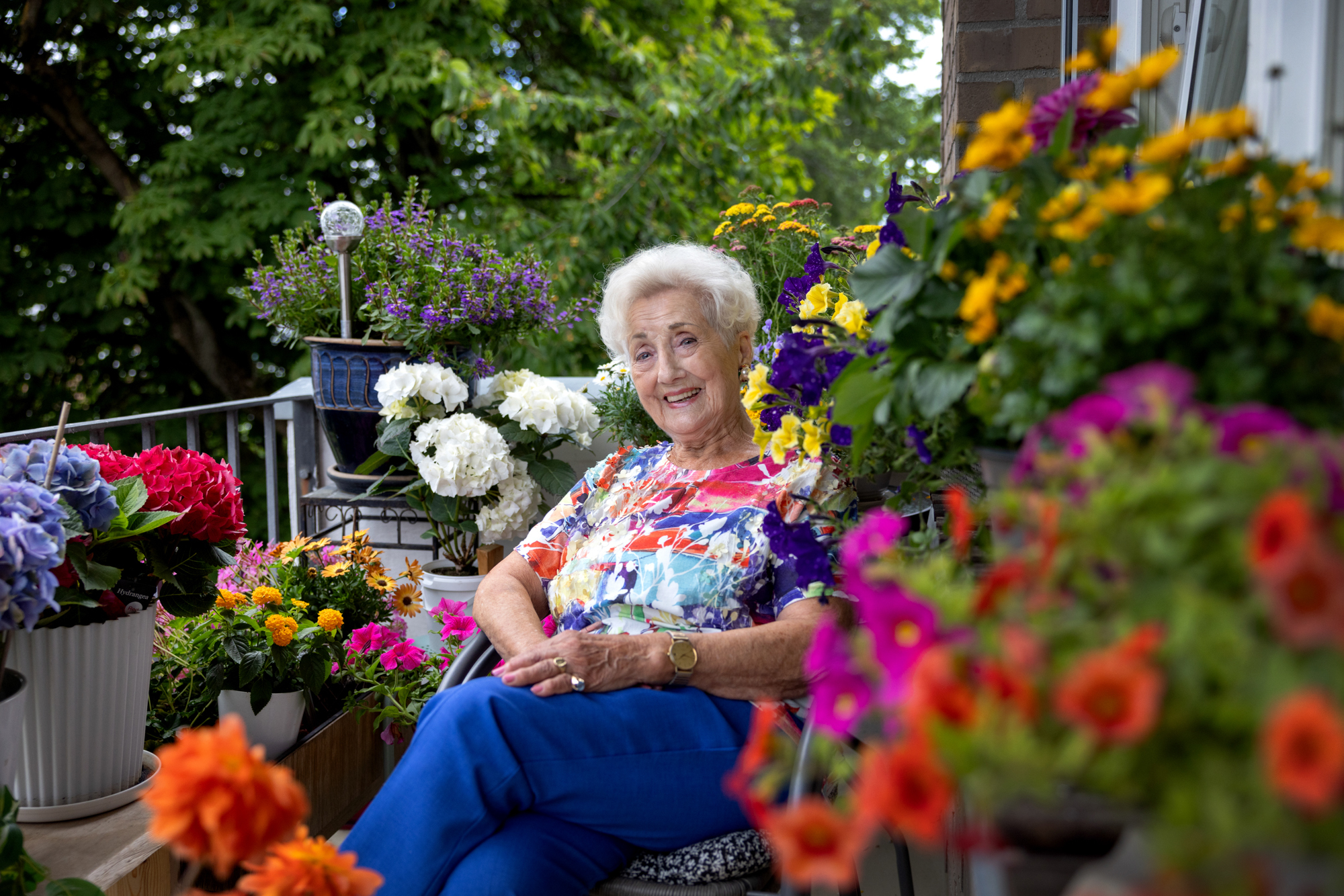 Foto: Kvinne sent i 80-årene med kort, grått hår smiler til fotografen. Hun sitter på balkongen omgitt av blomster i alle farger.