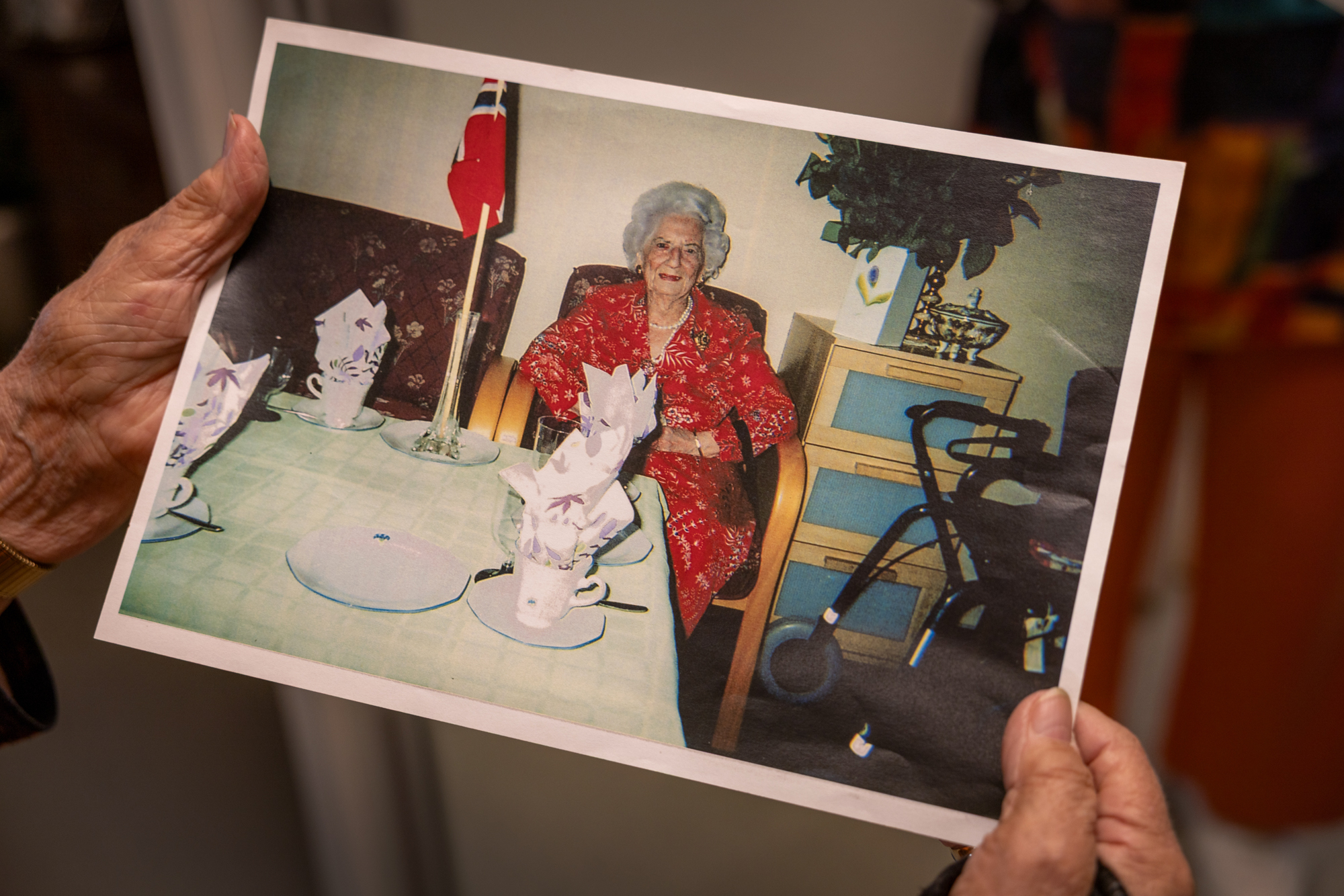 Foto: To gamle kvinnehender holder et bilde av en gammel dame på et rom foran et dekket bord med norsk flagg i en vase..
