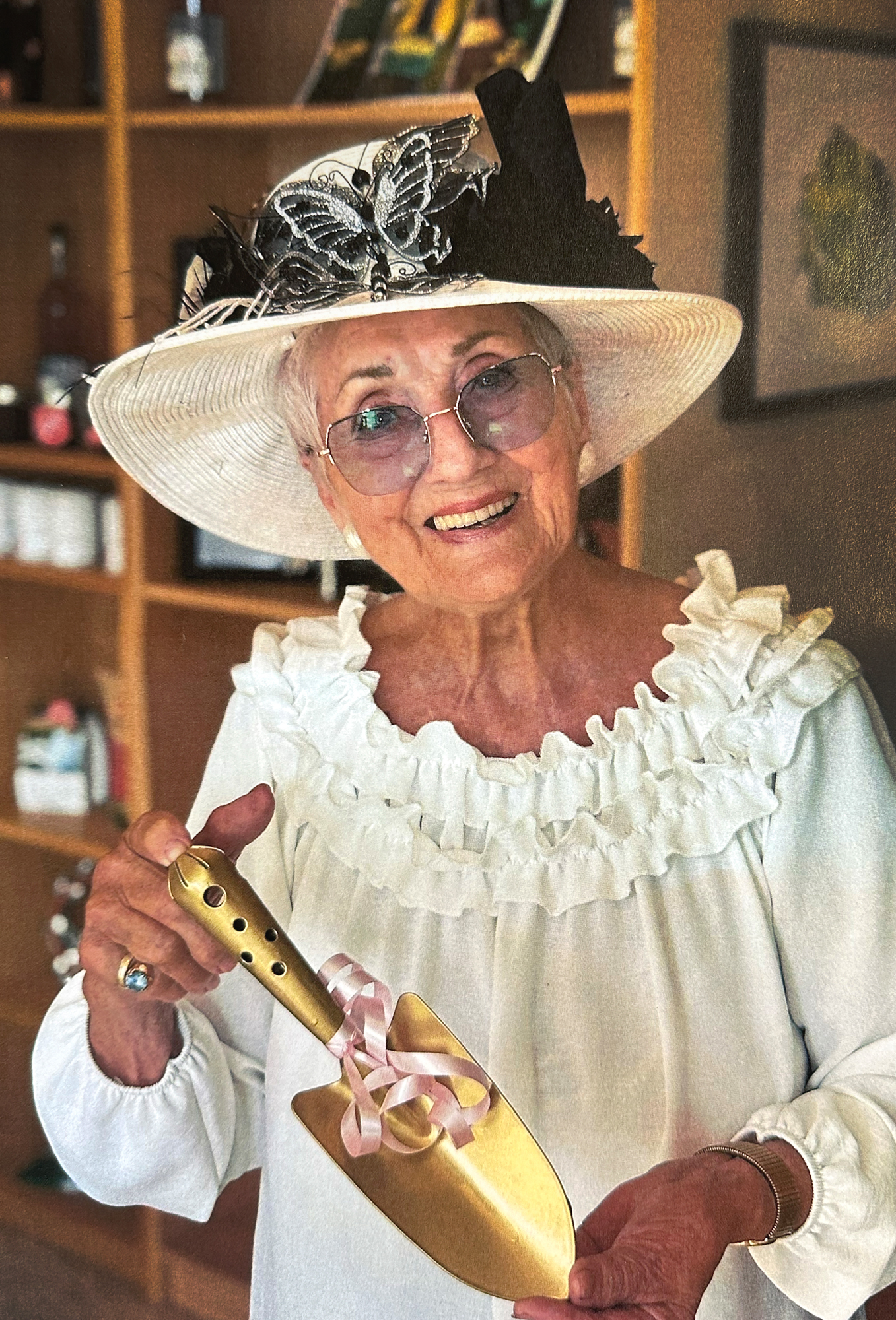 Foto: Gammel kvinne i hvit blose med rysjer rundt halsen og hvit hatt med svart dekor. Hun holder opp en hagespade som er gullforgylt. Den har rosa sløyfe på seg.
