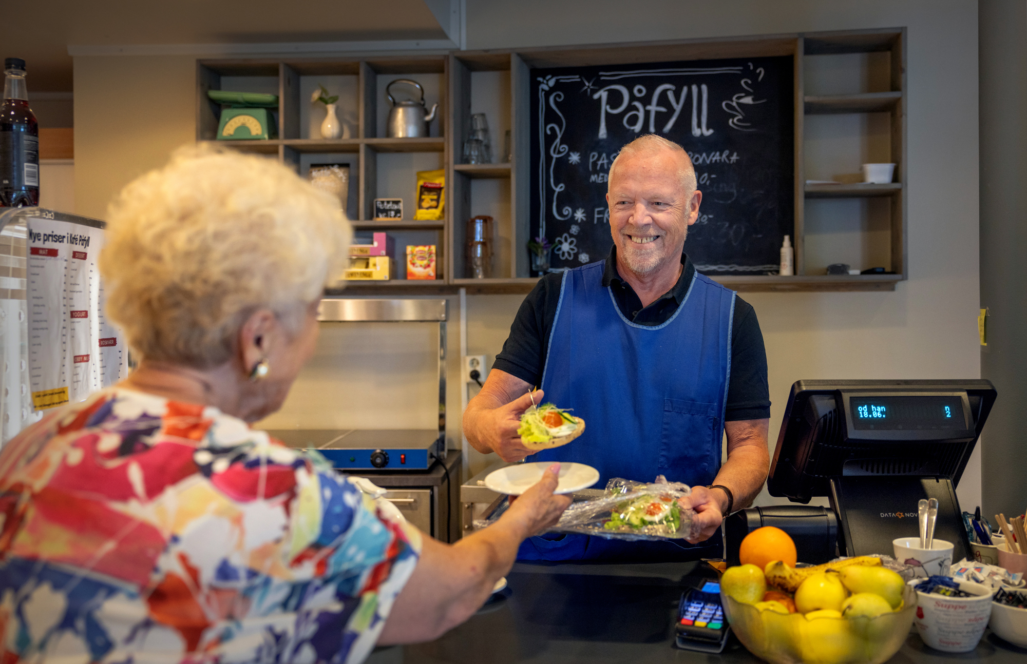 Foto: Kvinne i fargerik bluse med ryggen til får servert noe mat av en voksen mann med blått forkle i en kafe.