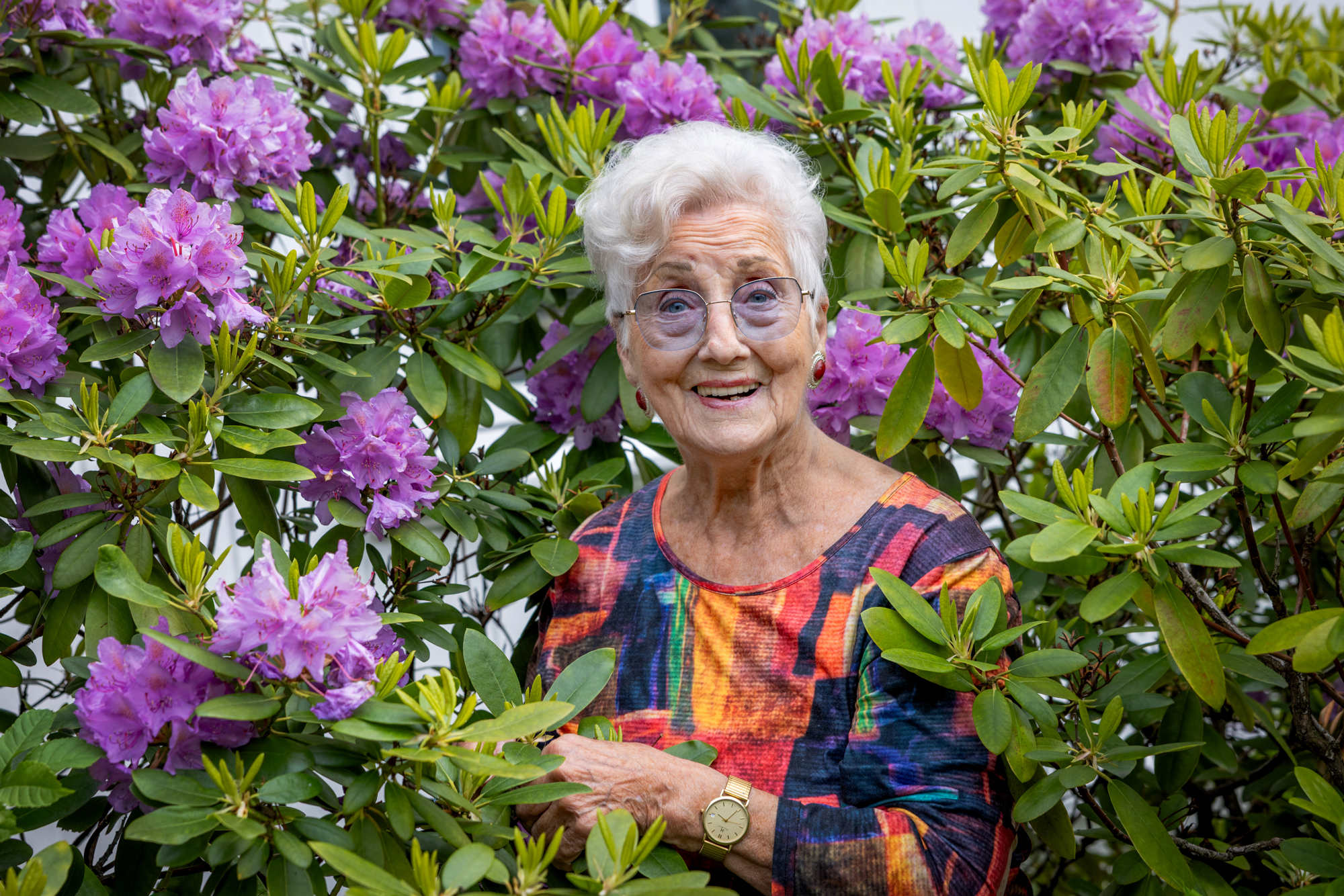 Foto: Eldre kvinne med kort, grått hår og briller med lilla glass stikker hodet fram blant lilla blomster.