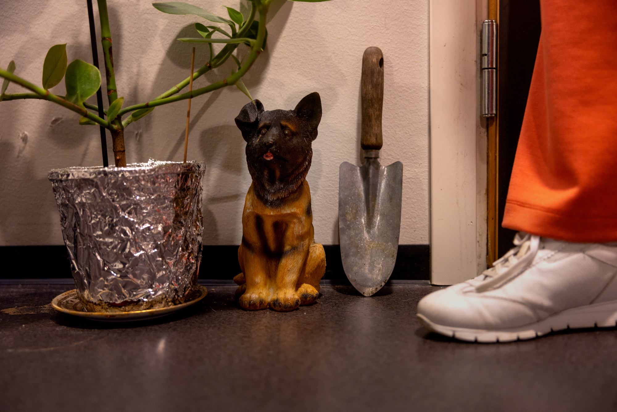 Foto: En fot med oransje bukse og hvite joggesko kommer ut av en dør. Der står det bl.a. oppstilt enn hagespade sammen med en figur av en liten hund og en potteplante.
