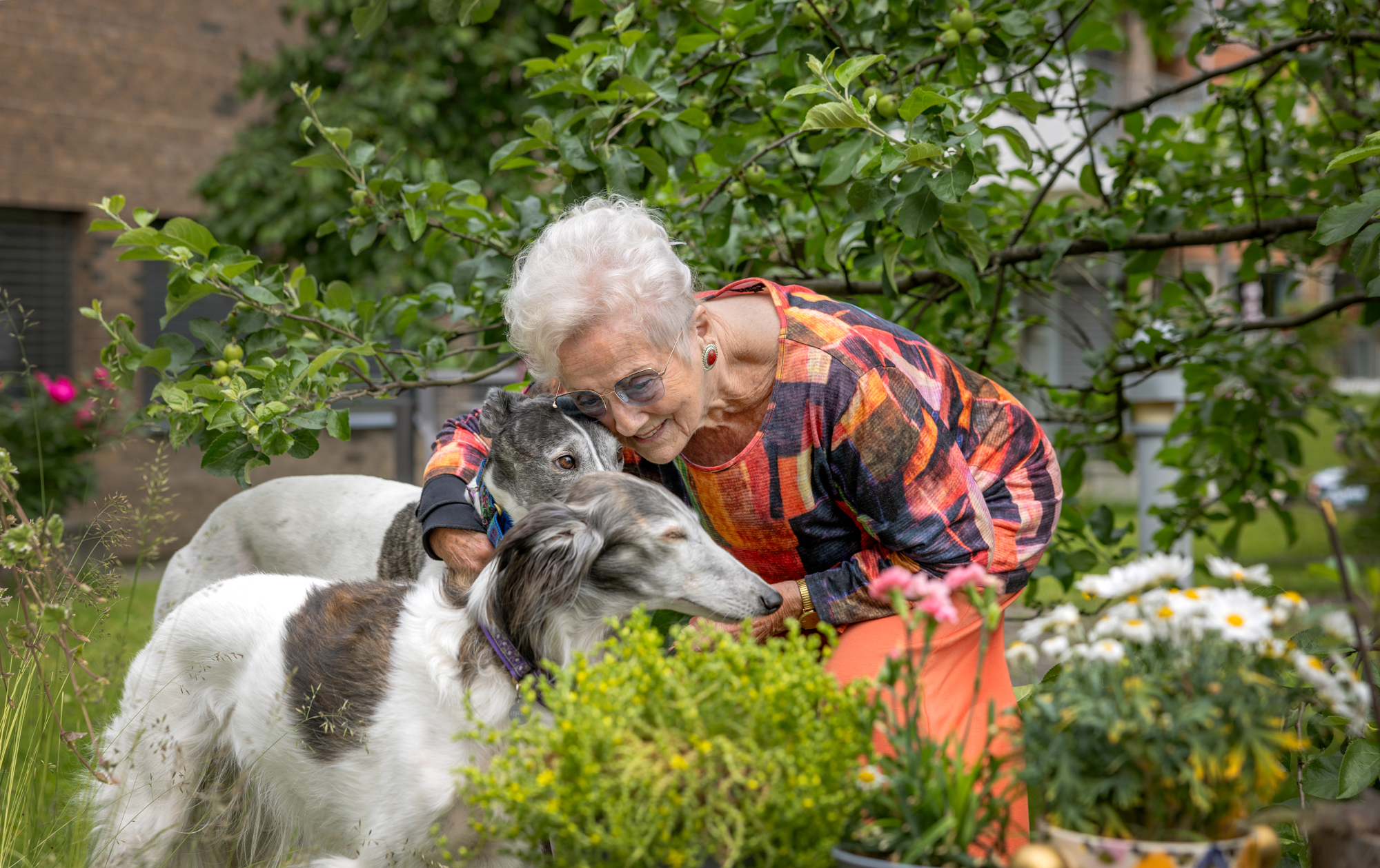 Foto: Eldre kvinne i mønstrrete topp og oransje bukser er i hagen og kose med to under.