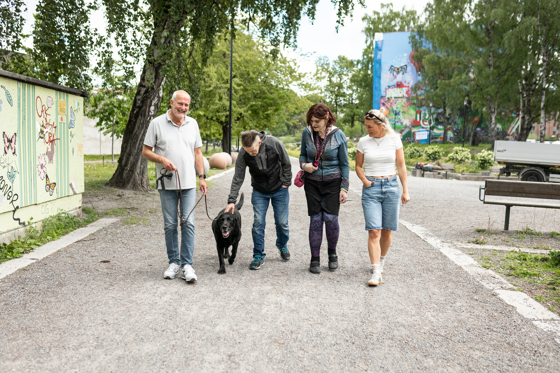 Foto: To menn og to kvinner går bortover en grusvei med trær og gress i bakgrunnen. De to mennene går til venstre, den ene holder den svarte labradoren i bånd, den andre lener seg over hunden og klapper ham på hodet.