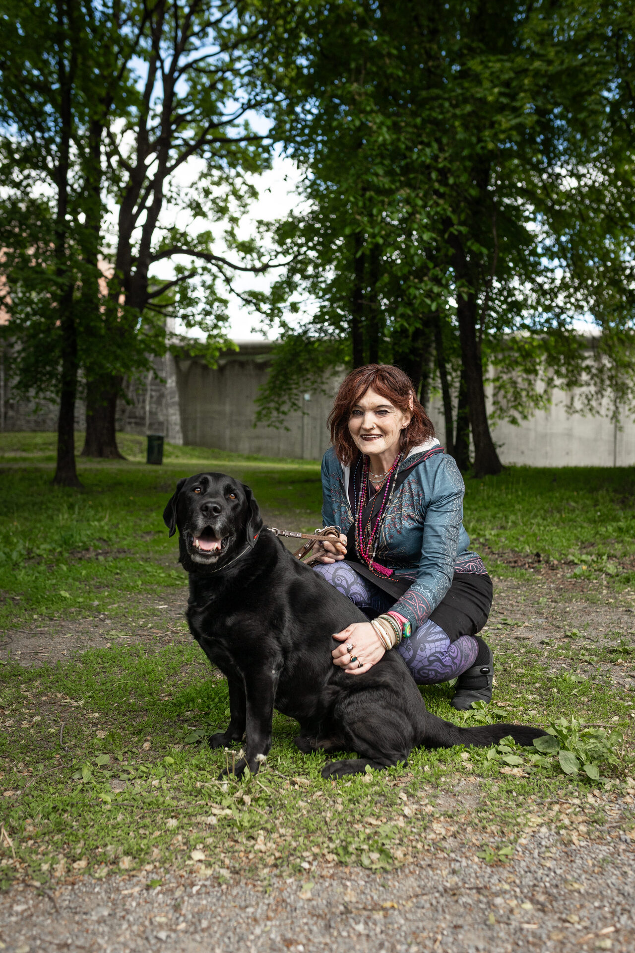 Foto: En kvinne sitter på huk i en park, og holder en svart labrador som sitter foran henne. Hun smiler mot kamera. Bak dem sees en høy, grå mur.