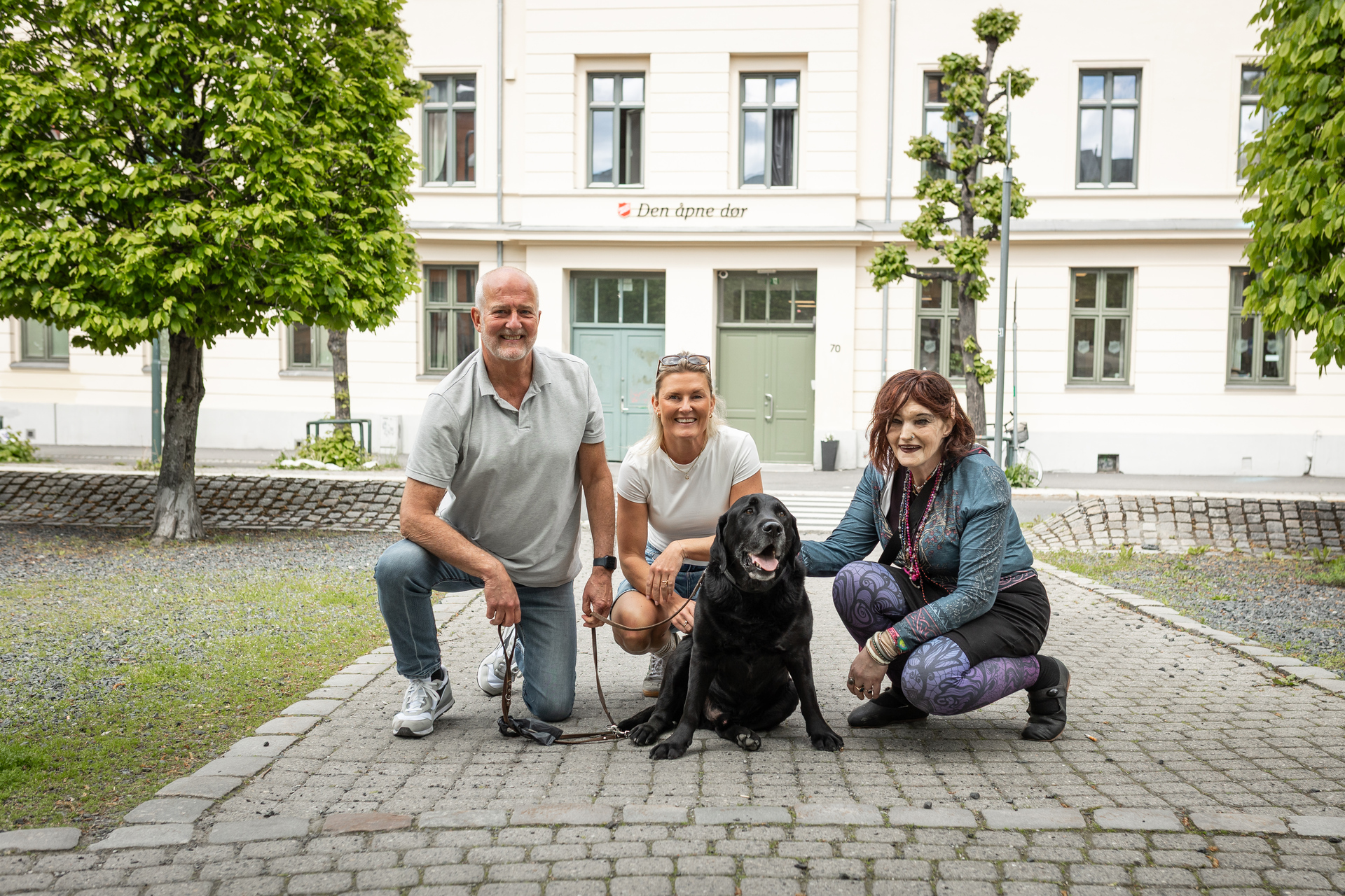 Foto: En mann, to kvinner og en hund med svart pels sitter på huk på brostein foran noen trær og et murbygg der det står Den åpne dør sammen med logoen til Frelsesarmeen.