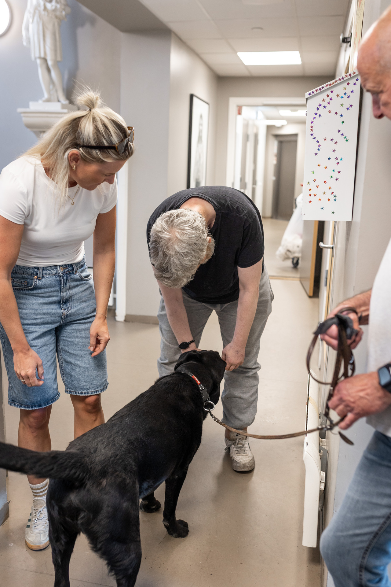 Foto: En mann med grått hår, svart t-skjorte og grå joggebukser står bøyd framover og koser med en svart labrador. Hunden står med ryggen til kamera. En kvinne står og smiler mot dem, og en mann står på den andre siden og holder i hundebåndet.