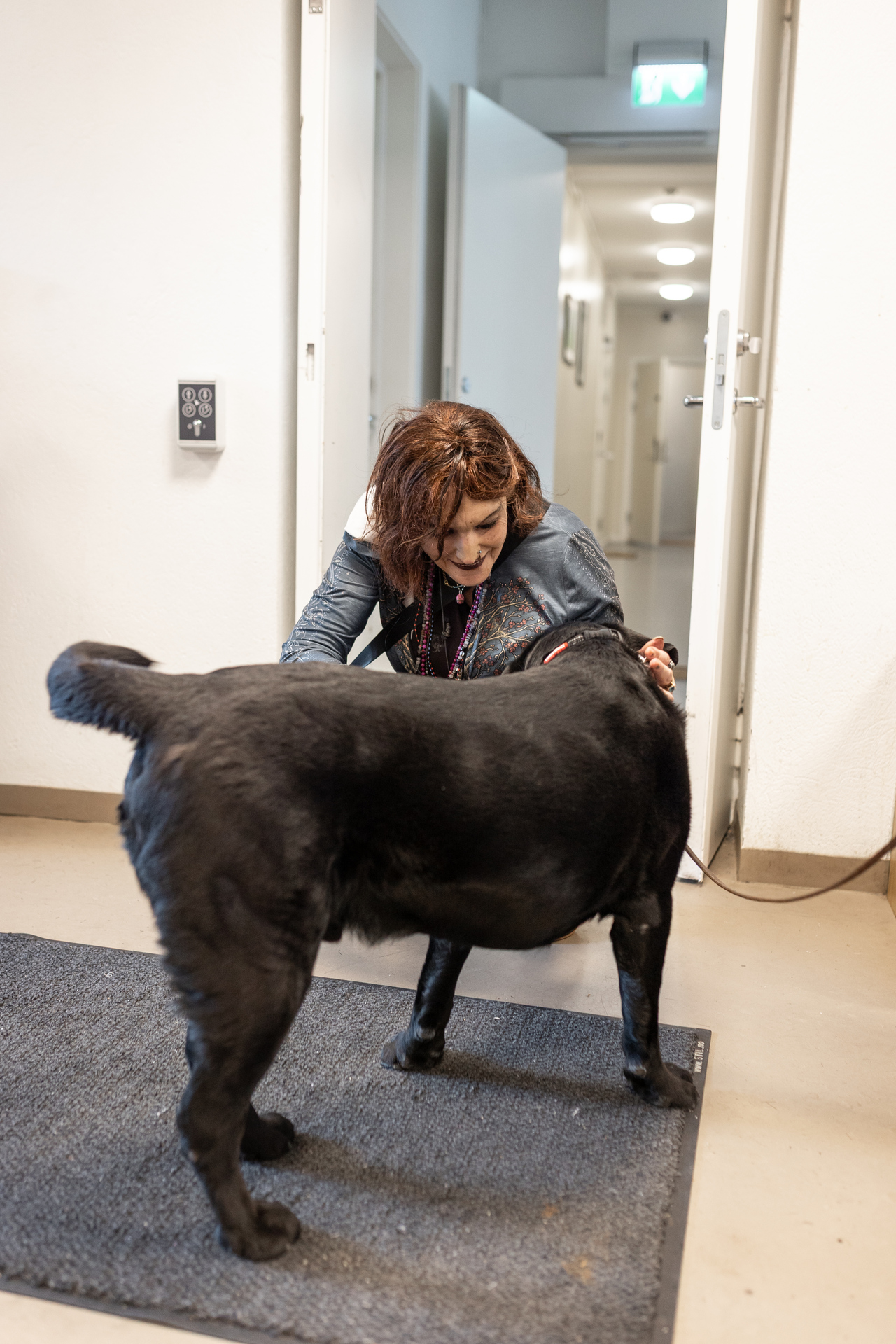 Foto: En dame med rødbrunt hår og olajakke sitter på huk og klapper en svart hund. Hun ser nedover mot hunden og smiler. Hunden står med ryggen til kamera og det ser ut som han logrer.