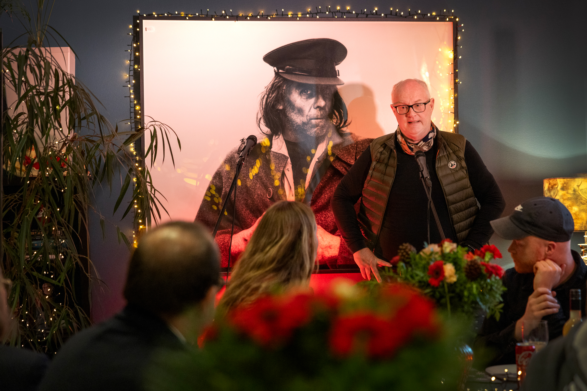 Eldre mann med briller som står foran på en scene. I bakgrunnen et stort bilde av en uteligger med hatt og frakk.