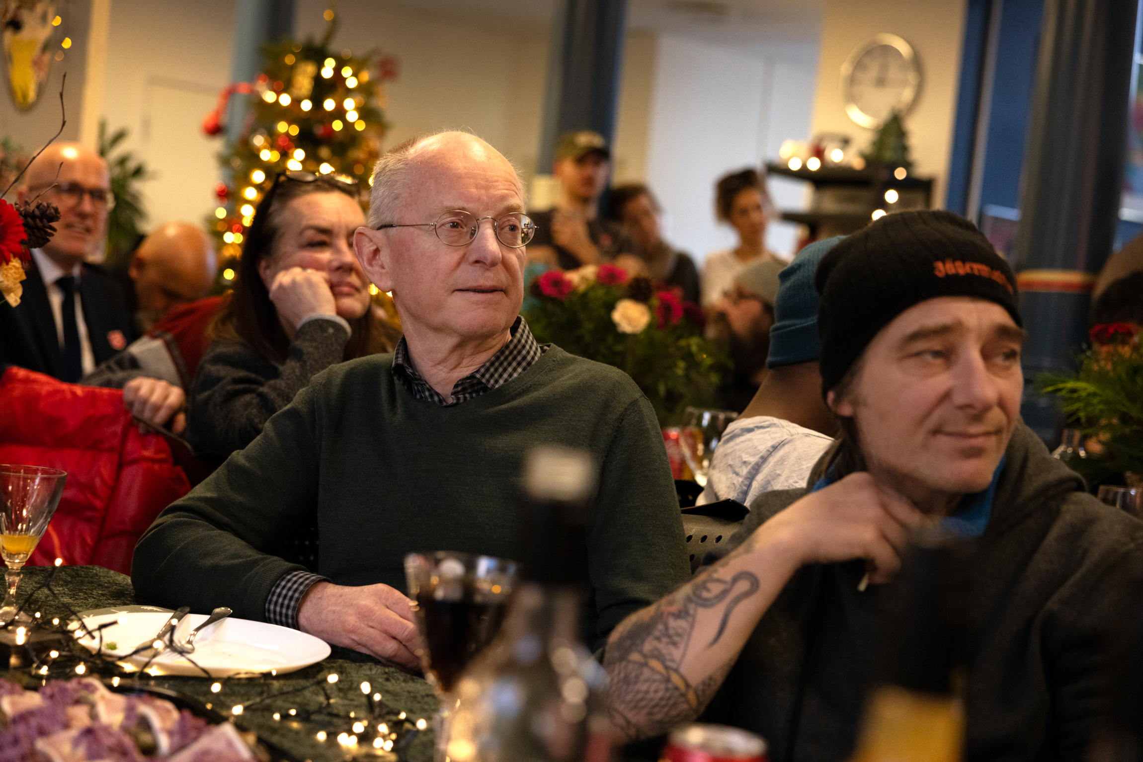 Et julepyntet lokale der folk sitter ved bordene. En eldre herre med briller sitter ved siden av en yngre mann med tatoveringer og lue.