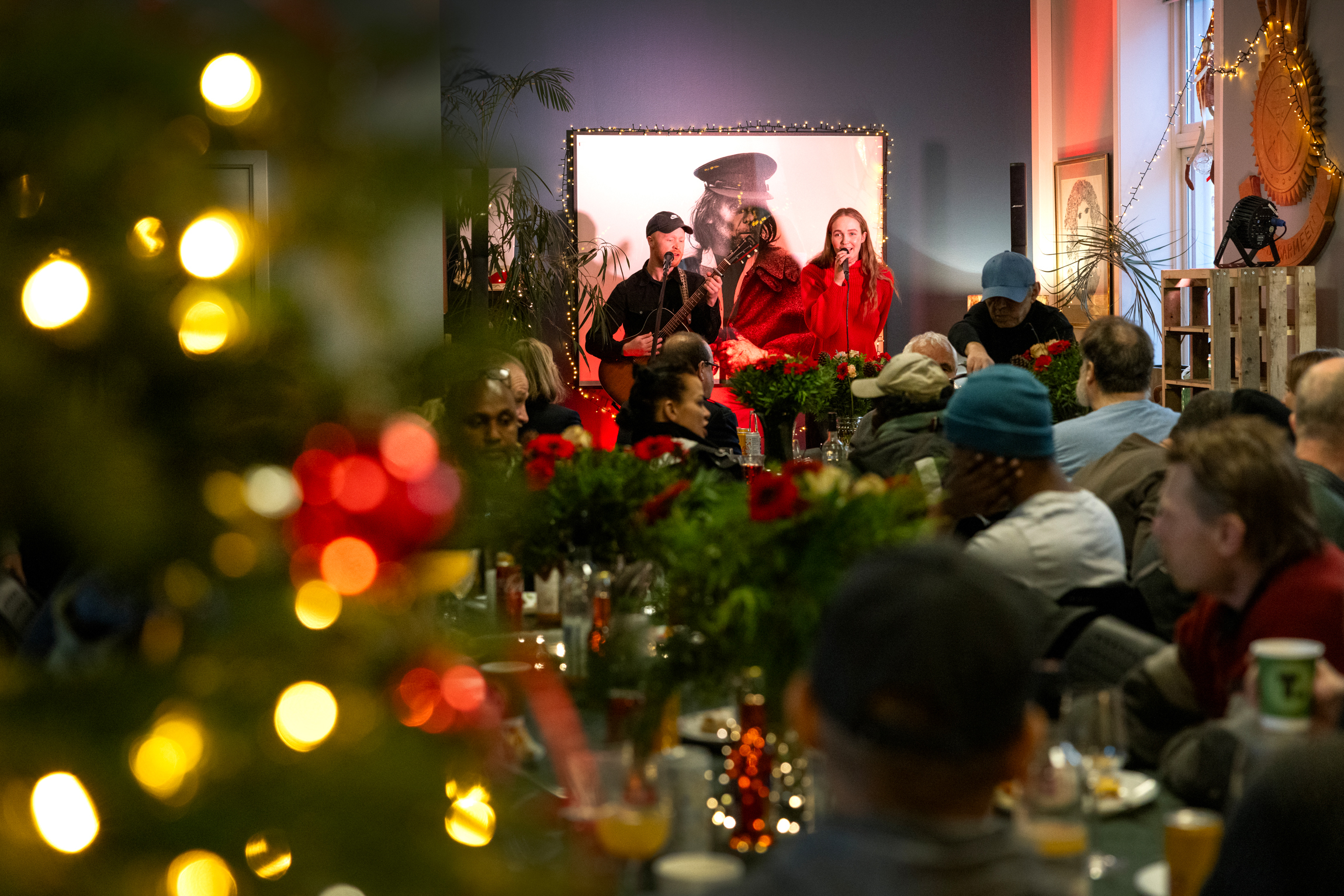 Unge kvinne med langt hår og rød genser synger fra scenen. Ved siden av står en gitarist. Lysene fra juletreet funkler på bildet.