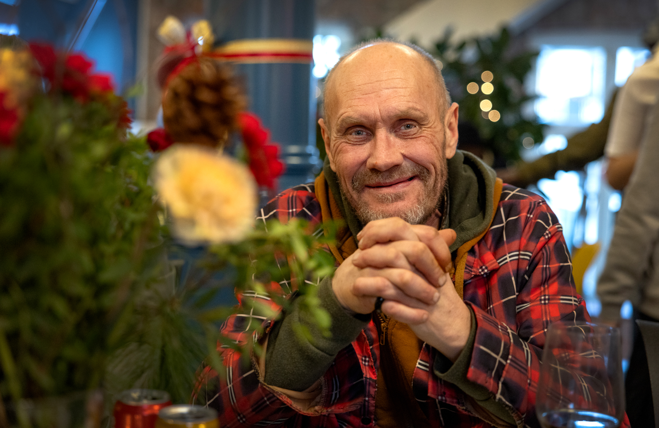 Mann sitter ved et bord og holder opp sine foldede hender. Han smiler og har på seg rutete ytterjakke inne i et julepyntet lokale.