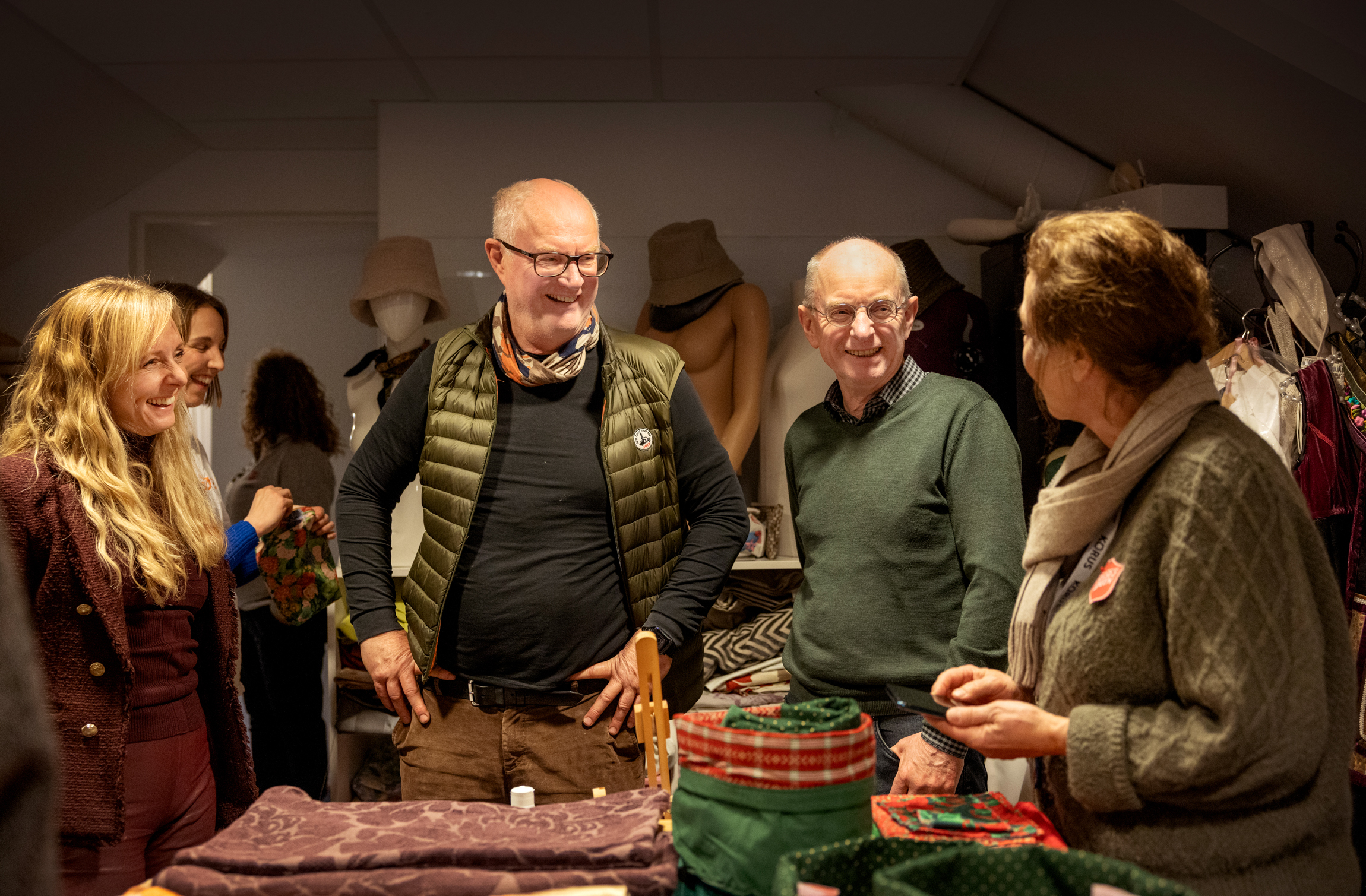Dame med langt, blondt hår står sammen med to eldre herrer. På et bord ved siden av dem ligger noen putetrekk som akkurat er sydd.