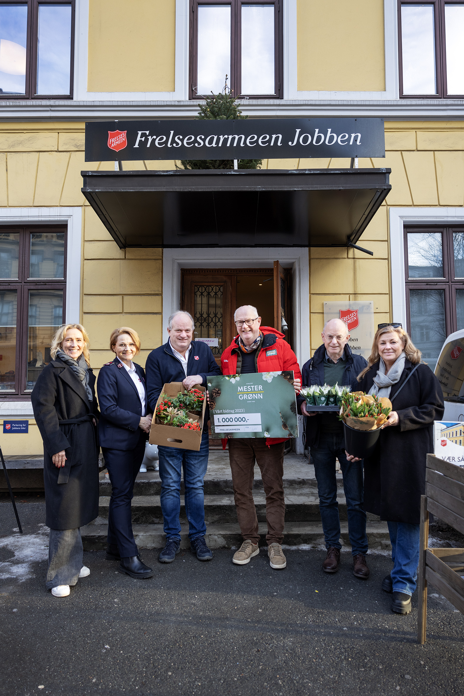 Tre kvinner og tre menn står utendørs i varme klær og holder esker med blomster og en diger papirsjekk. De står foran murhuset der Frelsesarmeen driver tiltaket Jobben Oslo.