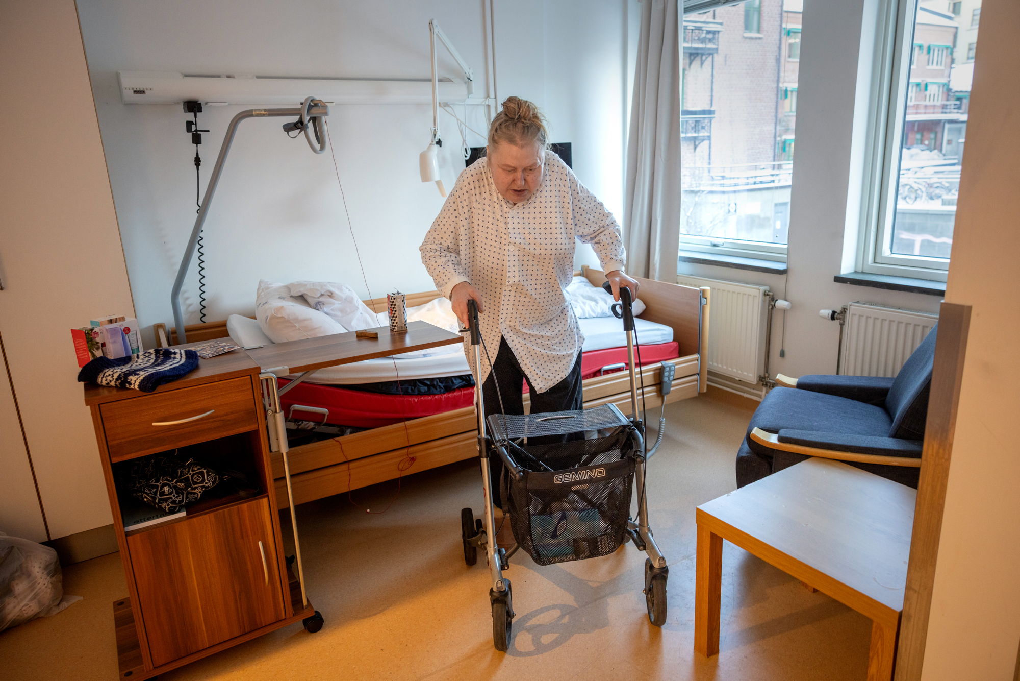 Foto: Kvinne i 50-årene har reist seg fra en sykehusseng og er på vei bortover et gulv med rullatoren sin. Hun har på seg svarte bukser og en hvit sykehusskjorte med mønster på. o
