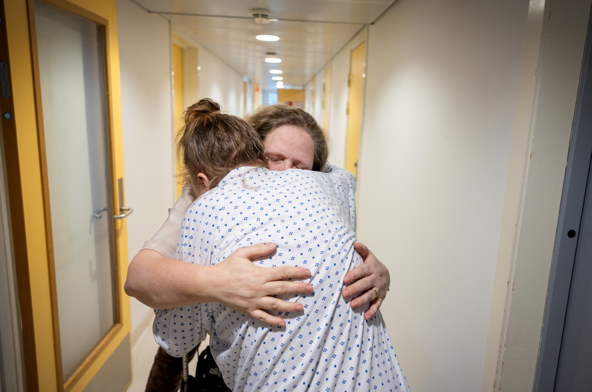 Foto: To middelaldrende kvinner klemmer hverandre i en sykehuskorridor. HUn med ryggen til har på seg sykehustrøye, den andre har på seg penkjær.