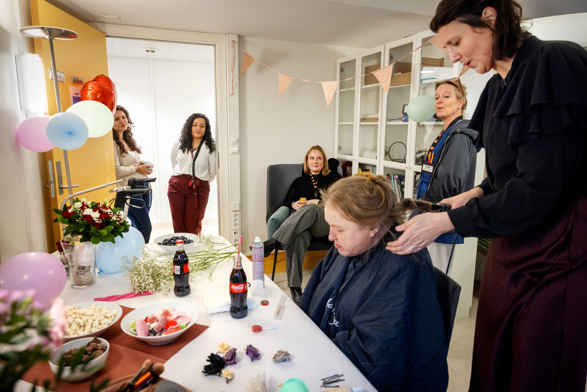 Foto: En yngre kvinne med mørkt hår bruker en krølltang i den mørkeblonde håret til en dame som sitter på en stol. På bordet foran damen står to flasker med Coca-Cola, godteri og annen snacks. Rundt står fire kvinner og ser på.