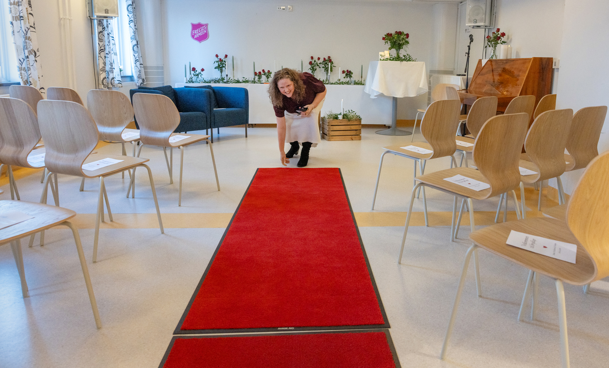 Foto: Dame med langt, krøllete hår og burgunder bluse justerer noen røde tepper i et rom med oppstilte stoler. I enden av rommet henger Frelsesarmeens logo på veggen.