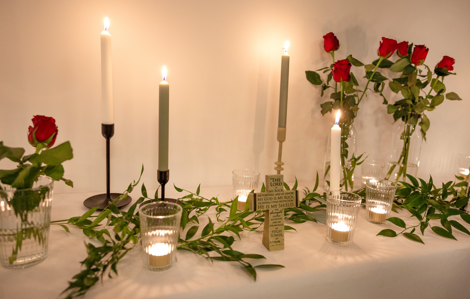 Foto: Smalt bord med hvit, lang duk står inntil en hvit vegg. Oppå er det pyntet med lys og blomster. Midt på står et lite kors.