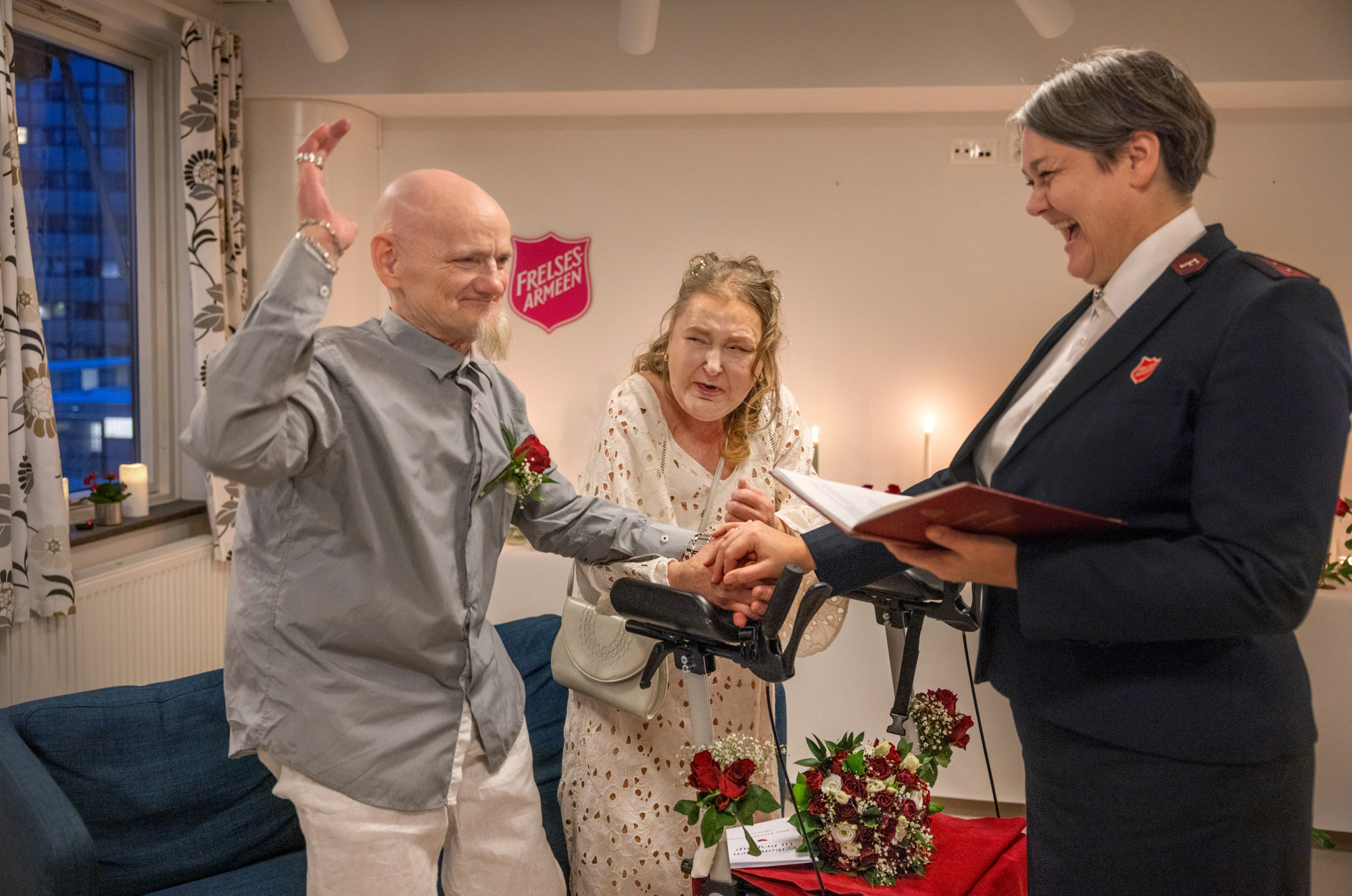 Foto: Middelaldrende kvinne med kort hår står i uniform fra Frelsesarmeen står foran et brudepar. Hun vier dem, og holder sin høyre hånd over deres hender. I den andre hånden holder hun en bok. Mannen og kvinnen som vies står bak en rullator, han uten hår med grått flippskjegg og hun i hvit kjole og nyfrisert hår. Brudebuketten ligger på setet til rullatoren.