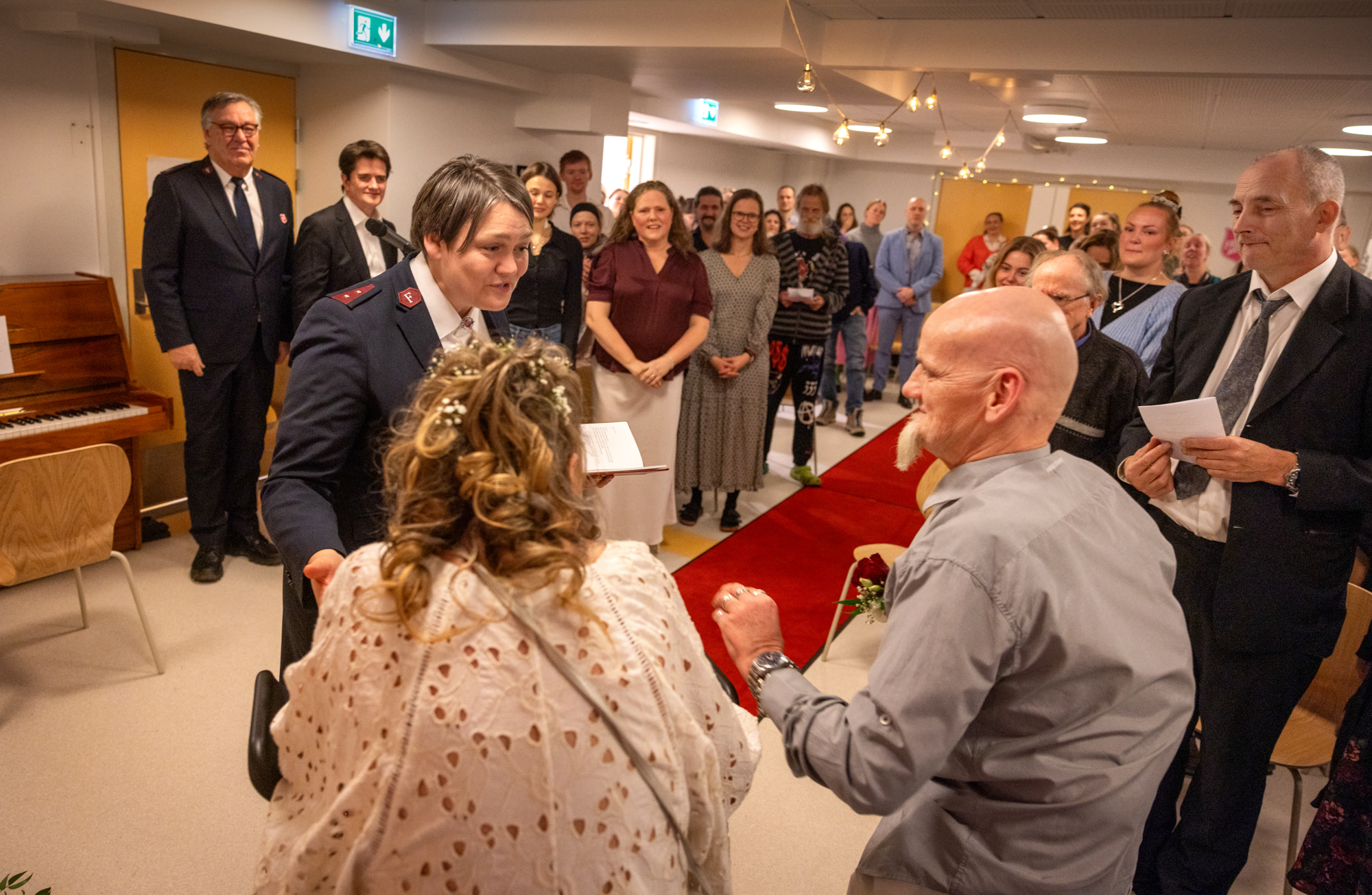 Foto: Vi ser brudeparet bakfra og ansiktet til kvinnen i uniform som vier de to. I det store rommet står det mange mennesker og ser på.