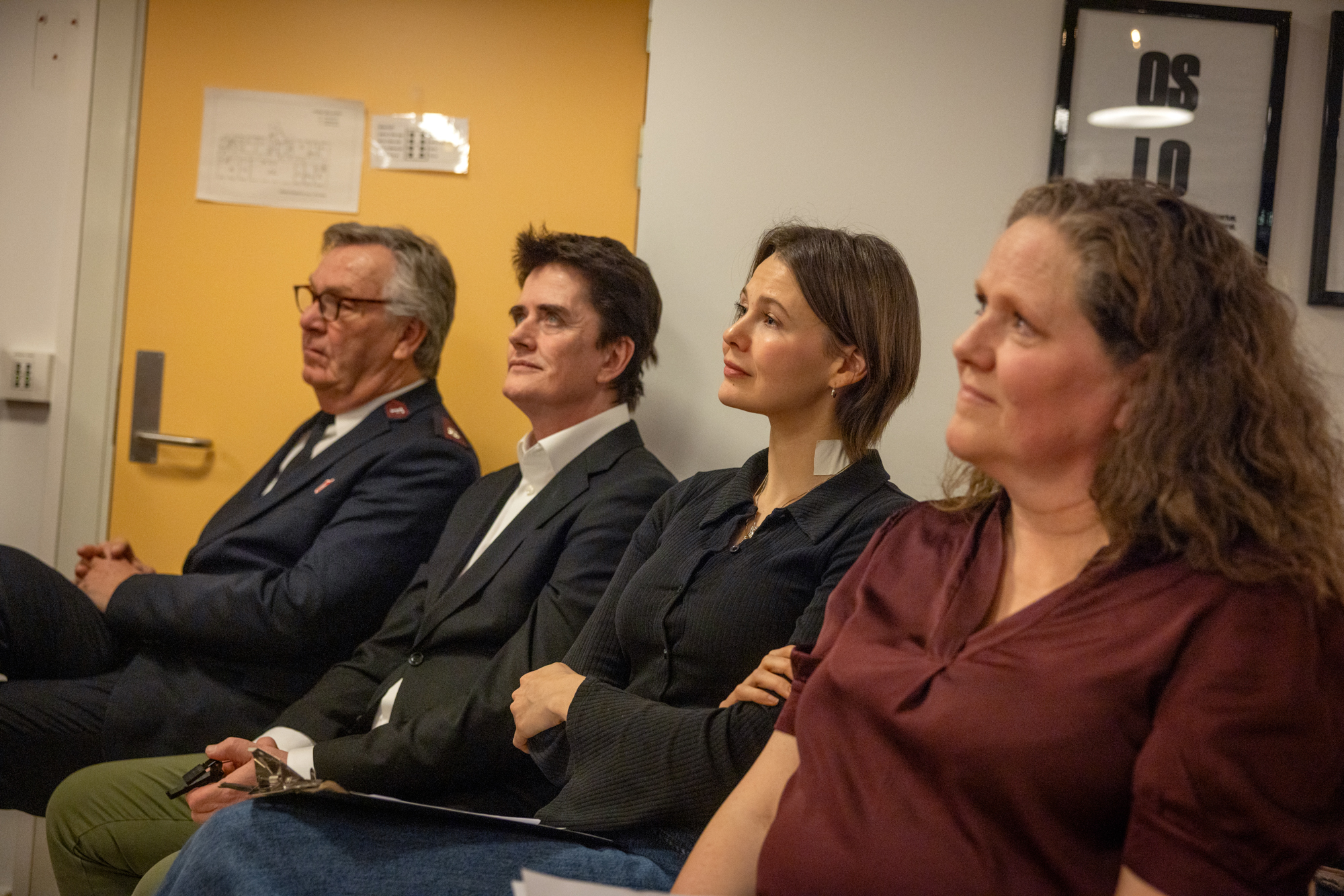 Foto: To menn og to kvinner sitter på rekke inntil en vegg. De ser alle mot samme punkt. Den ene mannen har uniform, de andre tre er pent kledd.