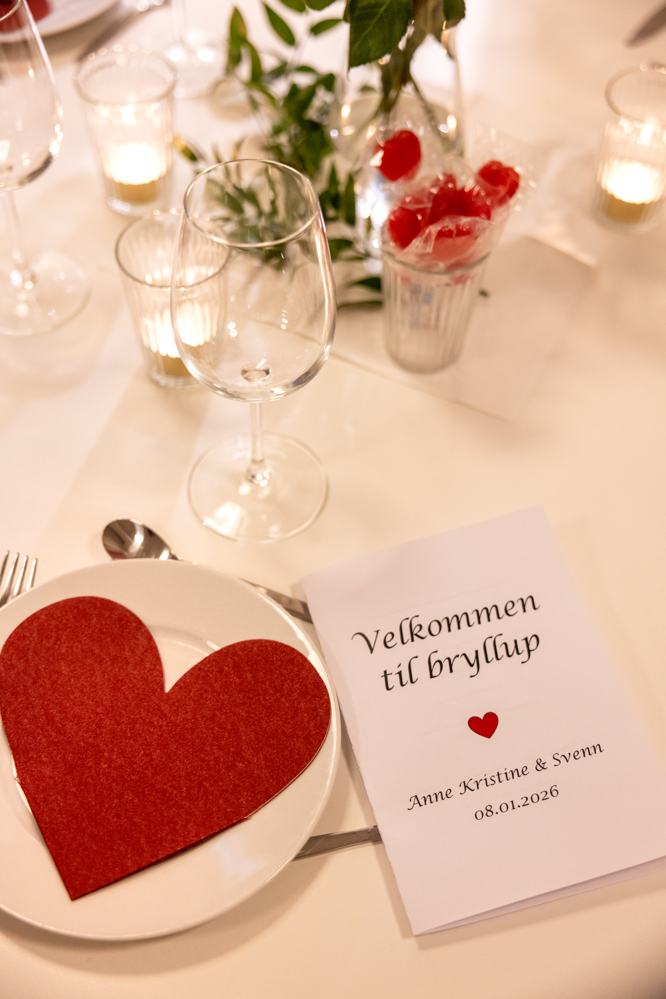 Foto: Dekket bord med glass, lys og røde filthjerter på tallerkener. Det ligger et lite program der det står Velkommen til bryllup, og så et rødt hjerte, to navn og dato nedover.