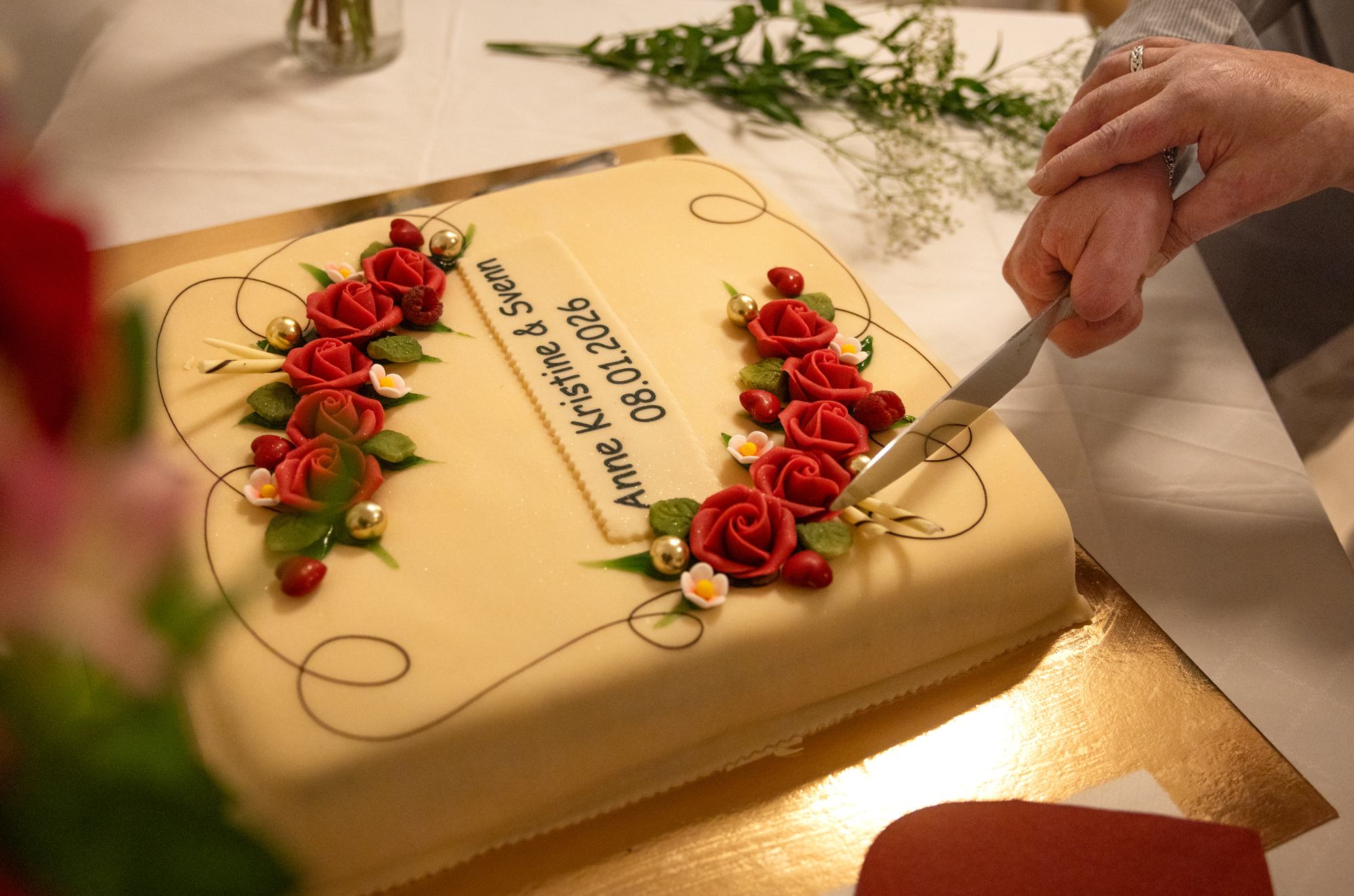 Foto: Hendene til en kvinne og mann holder i samme kakespade. De skjærer første stykke at en diger, firkantet marsipankake pyntet med røde roser.