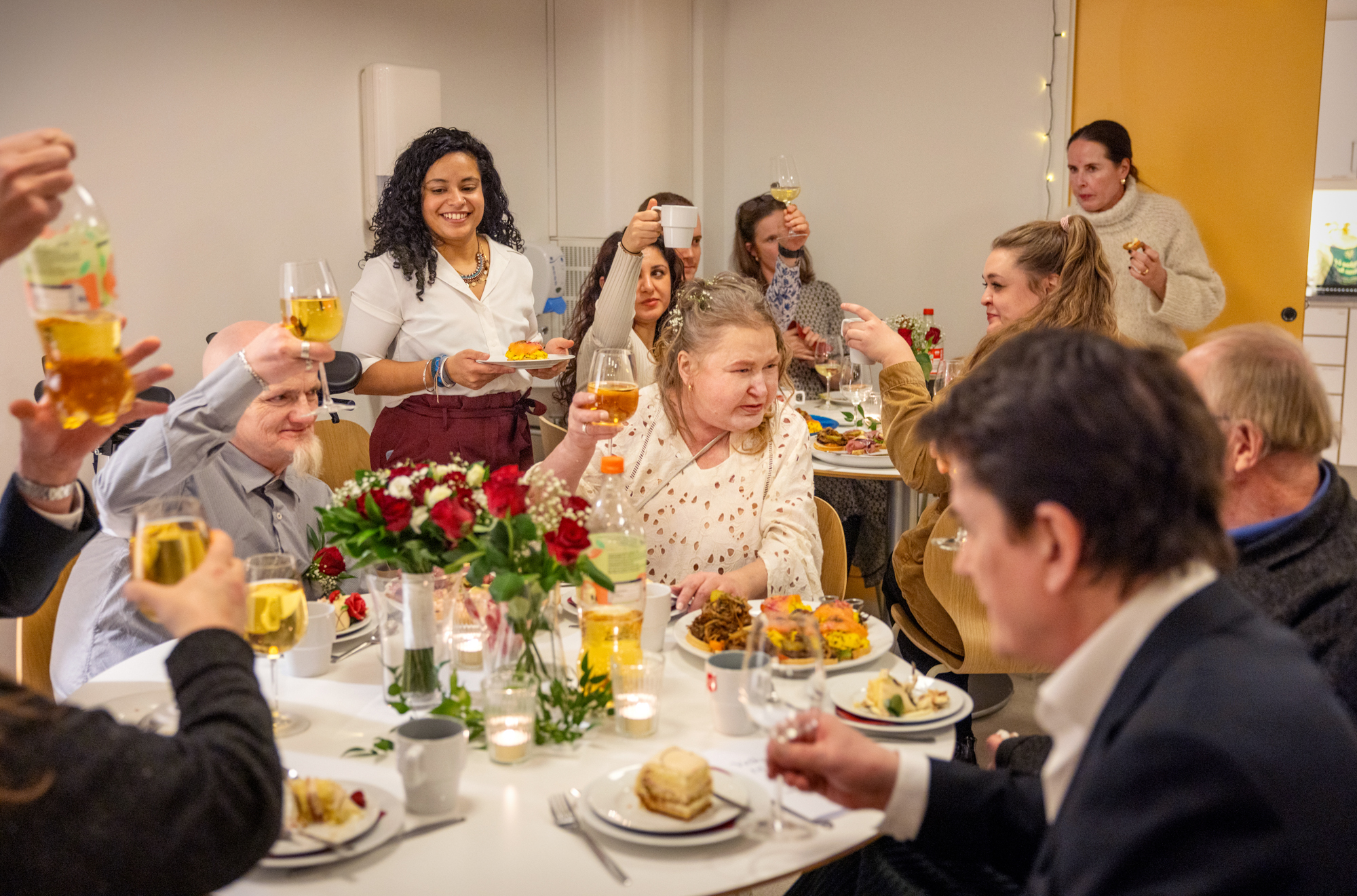 Foto: Flere bord fulle av glade mennesker som spiser og holde glasene høyt i en skål.