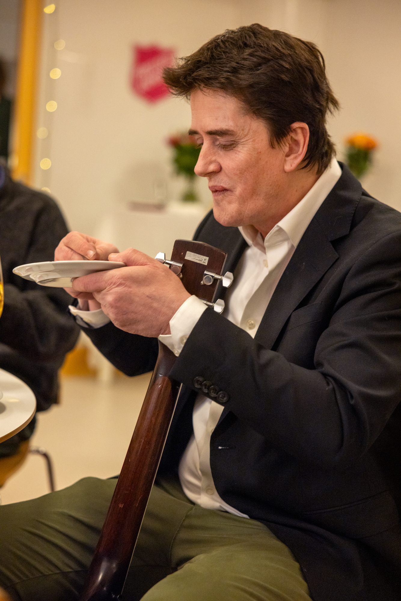 Foto: Mann med i mørk dressjakke holder en asjett opp mot munnen. Han spiser kake og har en gitar stående opp fra gulvet. Han klemmer den fast med lårene.
