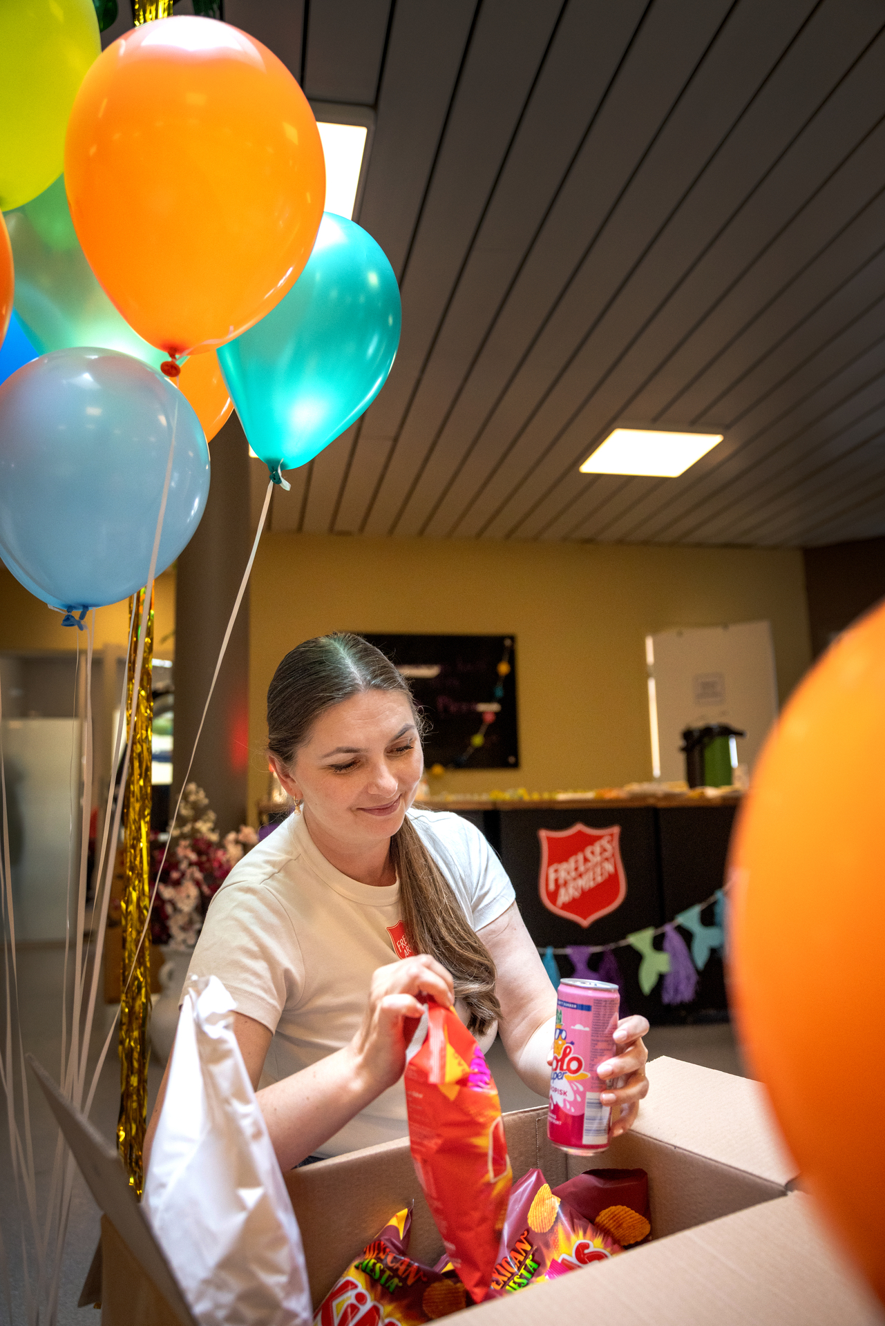 Foto: En kvinne med langt mørkt hår og hvit genser står og tar opp en brus og en pose potetgull av en kasse. Bak henne er mange heliumballonger.