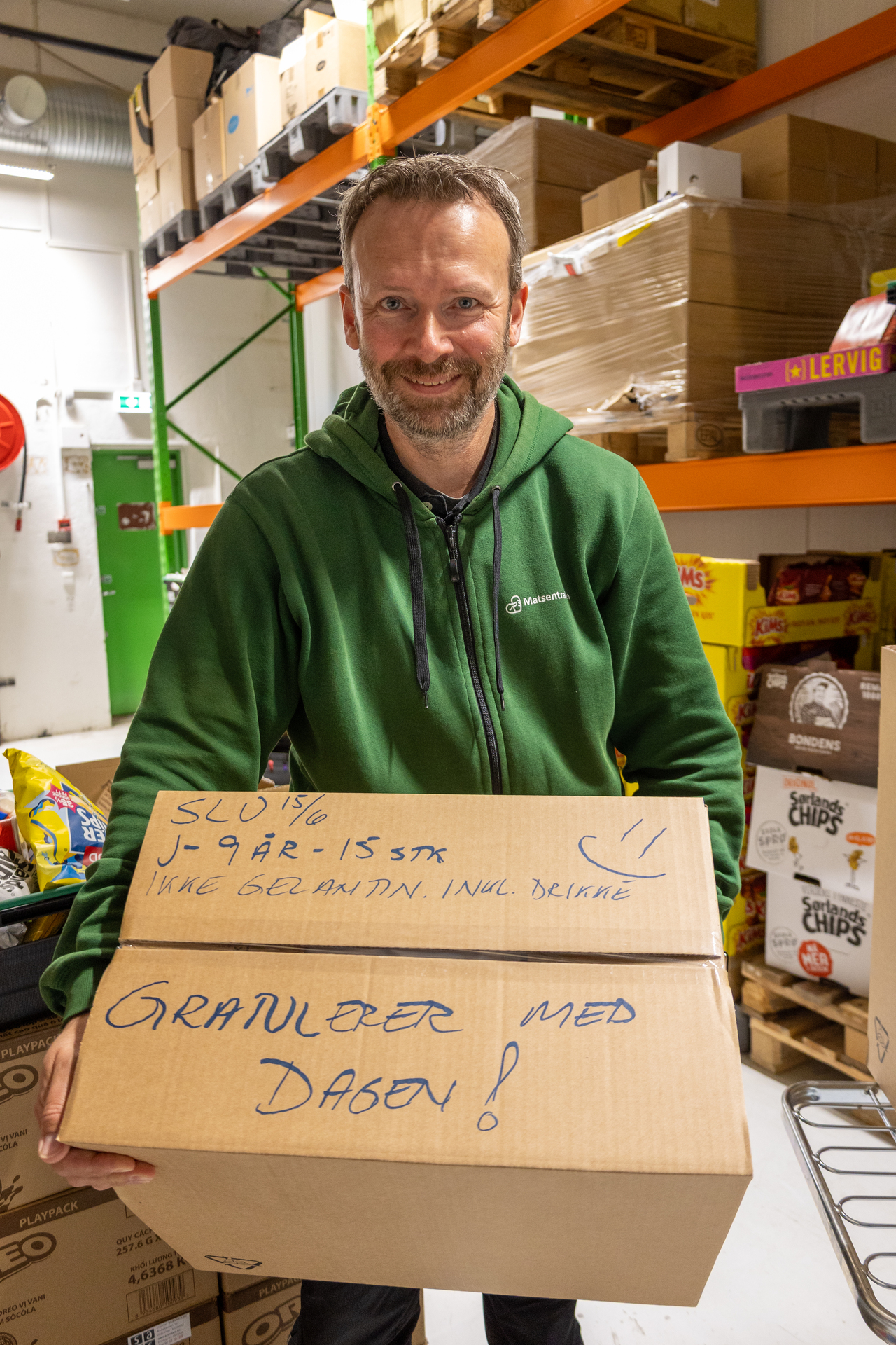 Foto: en mann med brunt, kort hår og kort skjegg og grønn hettegenser står og holder en stor pappkasse, med påskriften "Gratulerer med dagen!" Bak ham sees hyller med varer.