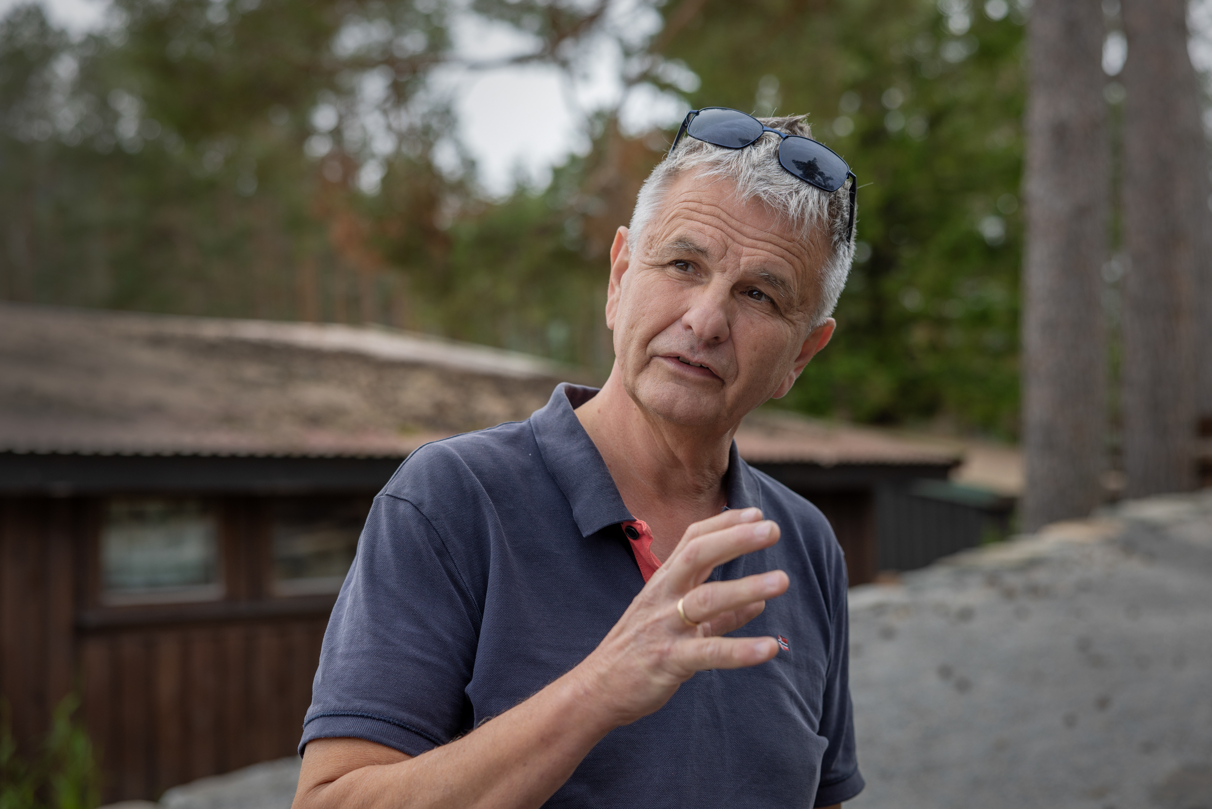 Foto: Mann i 60-årene som gestikulerer mens hans snakker. I bakgrunnen lavt, brunt bygg.