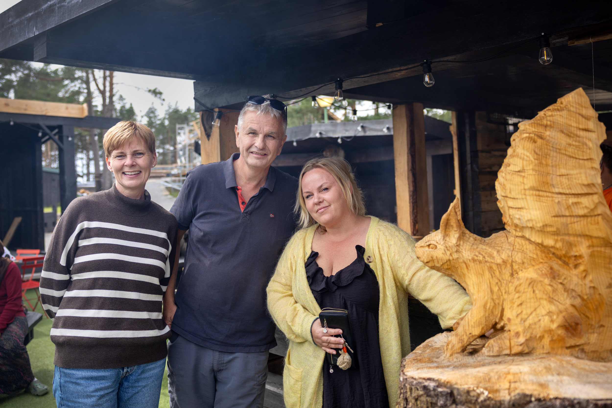 Foto:  Mann i 60-årene flankert av to damer i 40-årene. De står ute under tak, og det henger litt røyk fra en grill. På høyre side står en trestubbe med et utskåret ekorn på toppen.