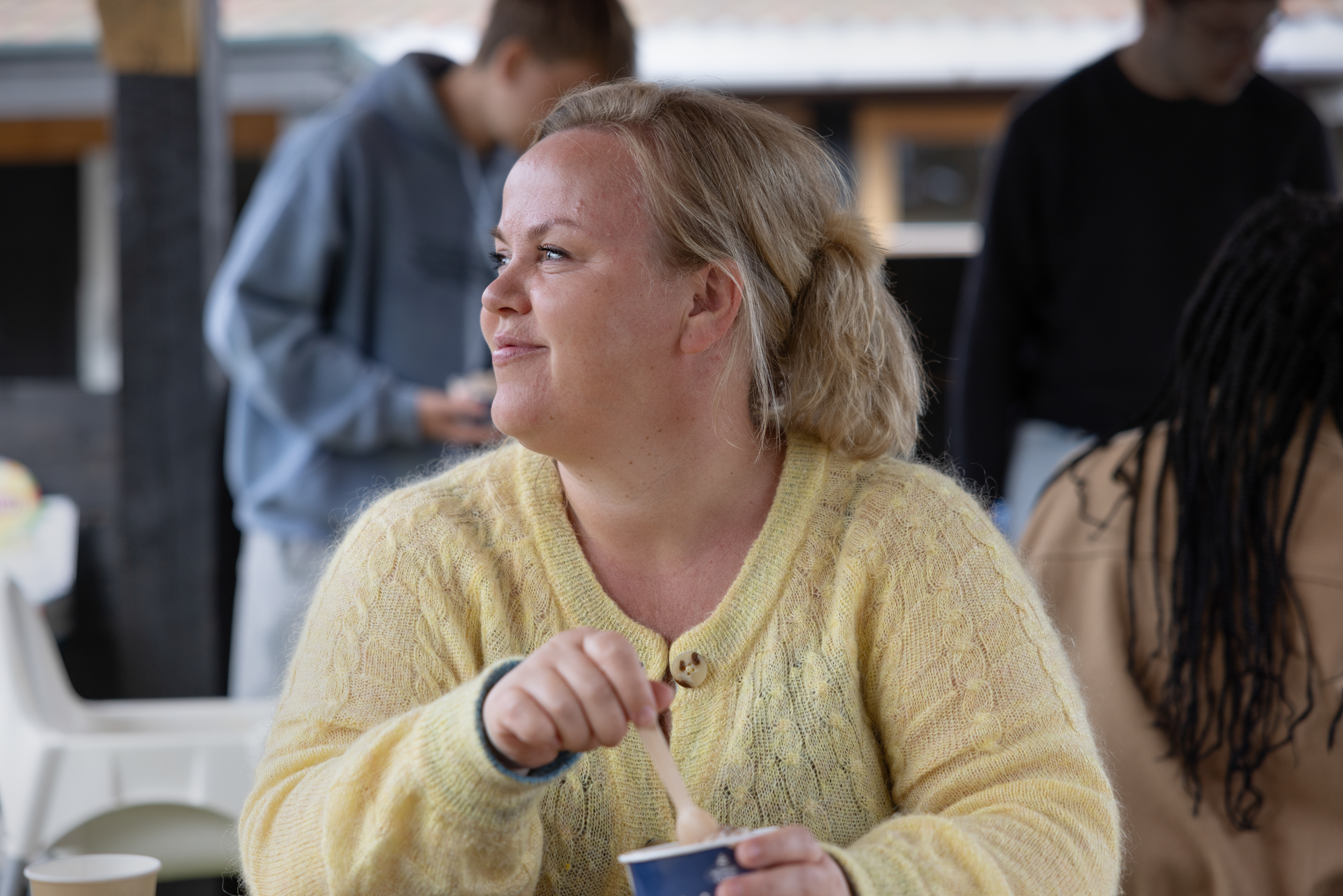 Foto: Kvinne med blondt hår spiser is og ser smilende bort. Rundet henne er det flere ungdommer.
