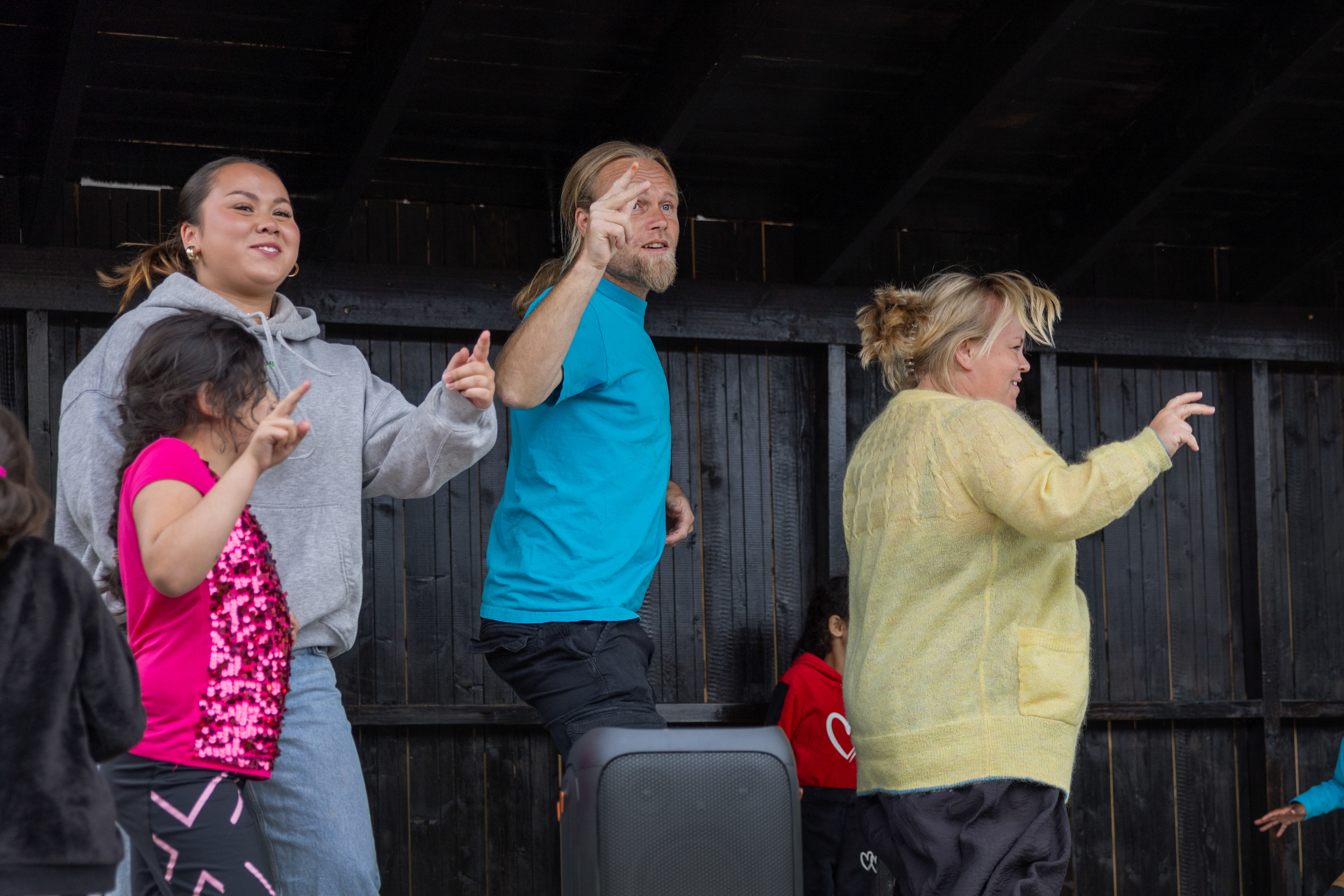Foto: En tenåringsjente i hettegenser, en blond mann med hestehale og T-skjorte en blond kvinne i tynn strikkejakke danser på scenen med flere barn rundt.