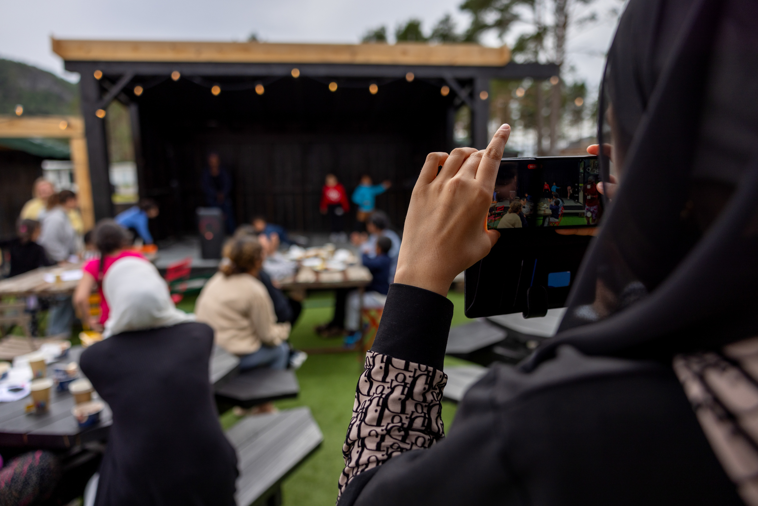 Foto: En ung dame med slør tar bilde av de som opptrer på scenen. Rundt sitter det masse folk på bord og spiser middag.