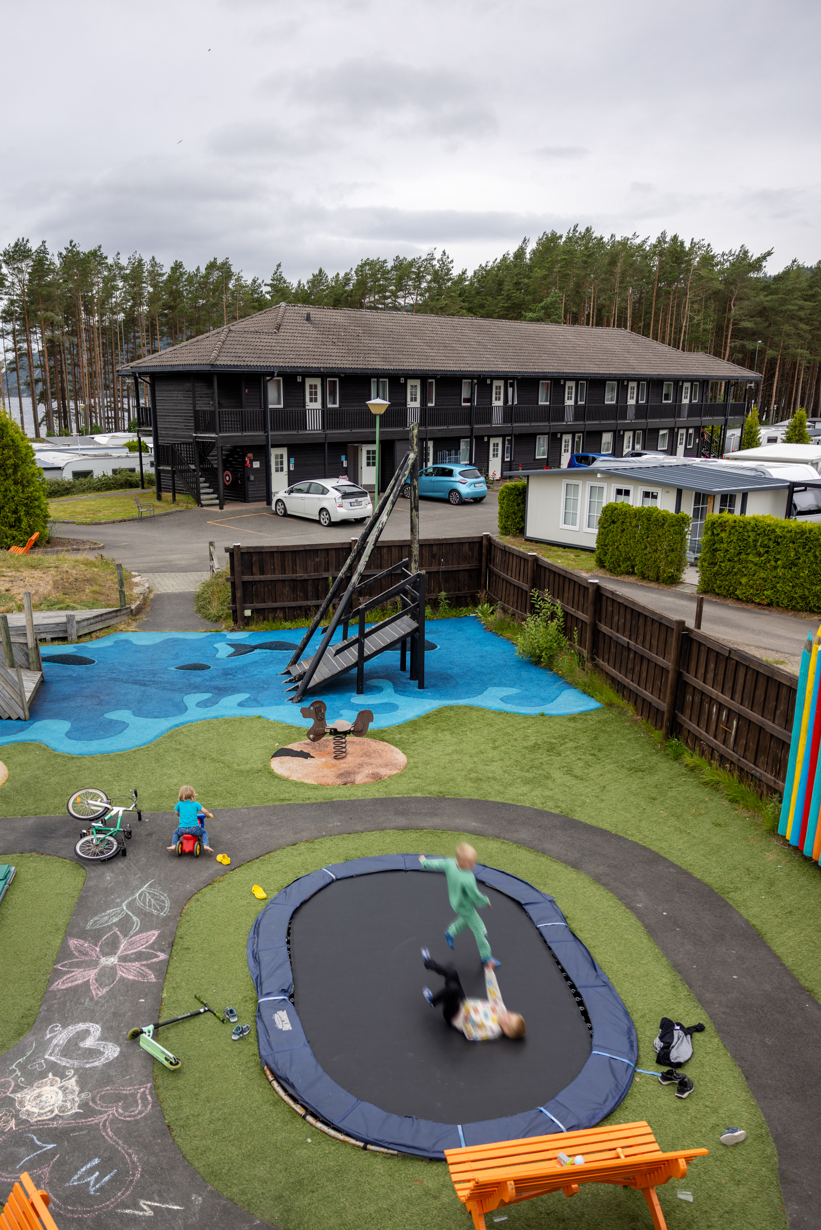 Foto: Oversiktsbilde av lekeplassen og et leilighetskompleks litt bortenfor. På begge sider står det campingvogner.