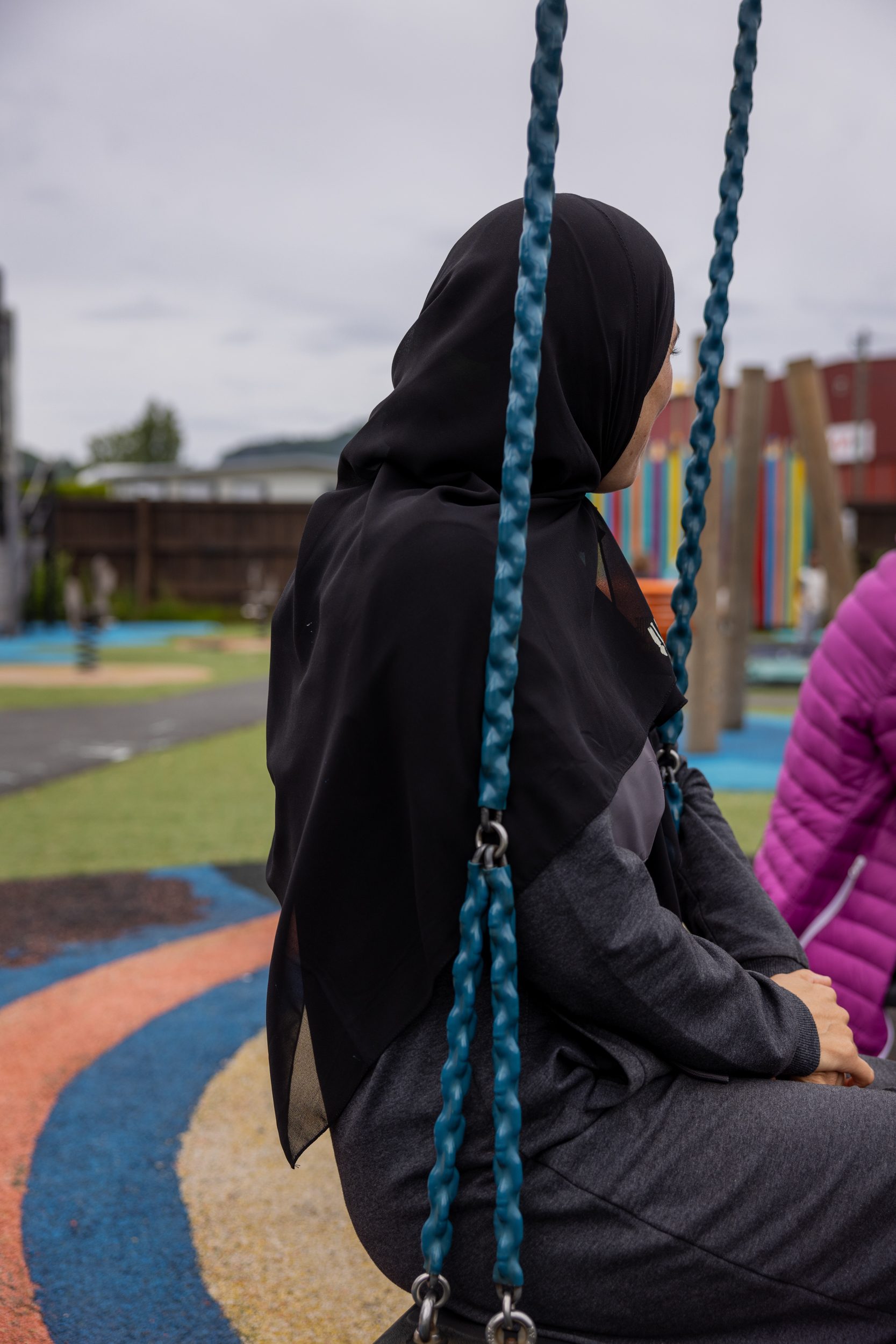 Foto: Kvinne med hijab sitter på en huske. På husken ved siden av ser vi litt av en rosa boblejakke.
