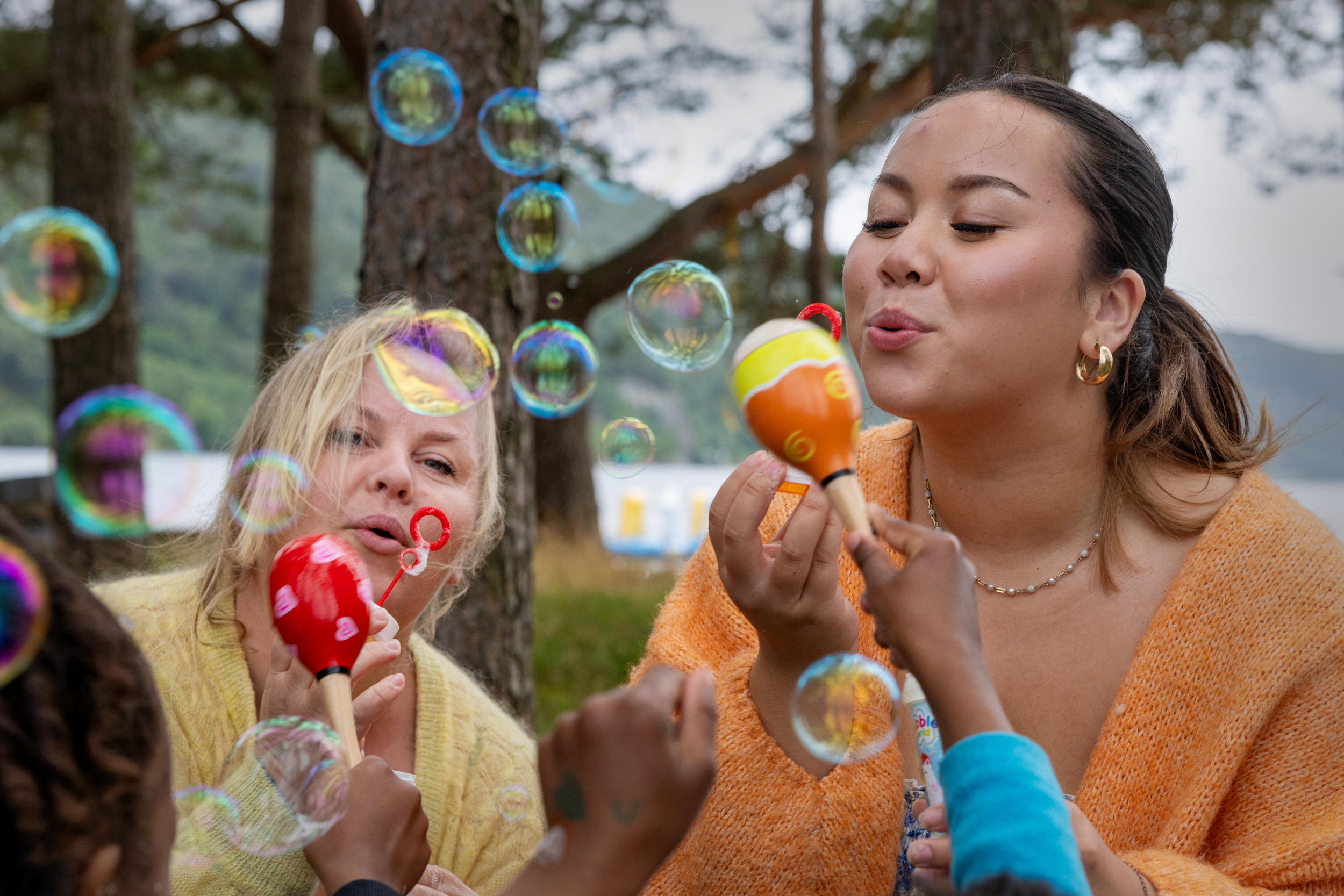 Foto: Blond kvinne i 40-årene og tenåringsjente med brunt hår blåse såpebobler. To barn med ryggen til strekker ut armene for å fange dem.