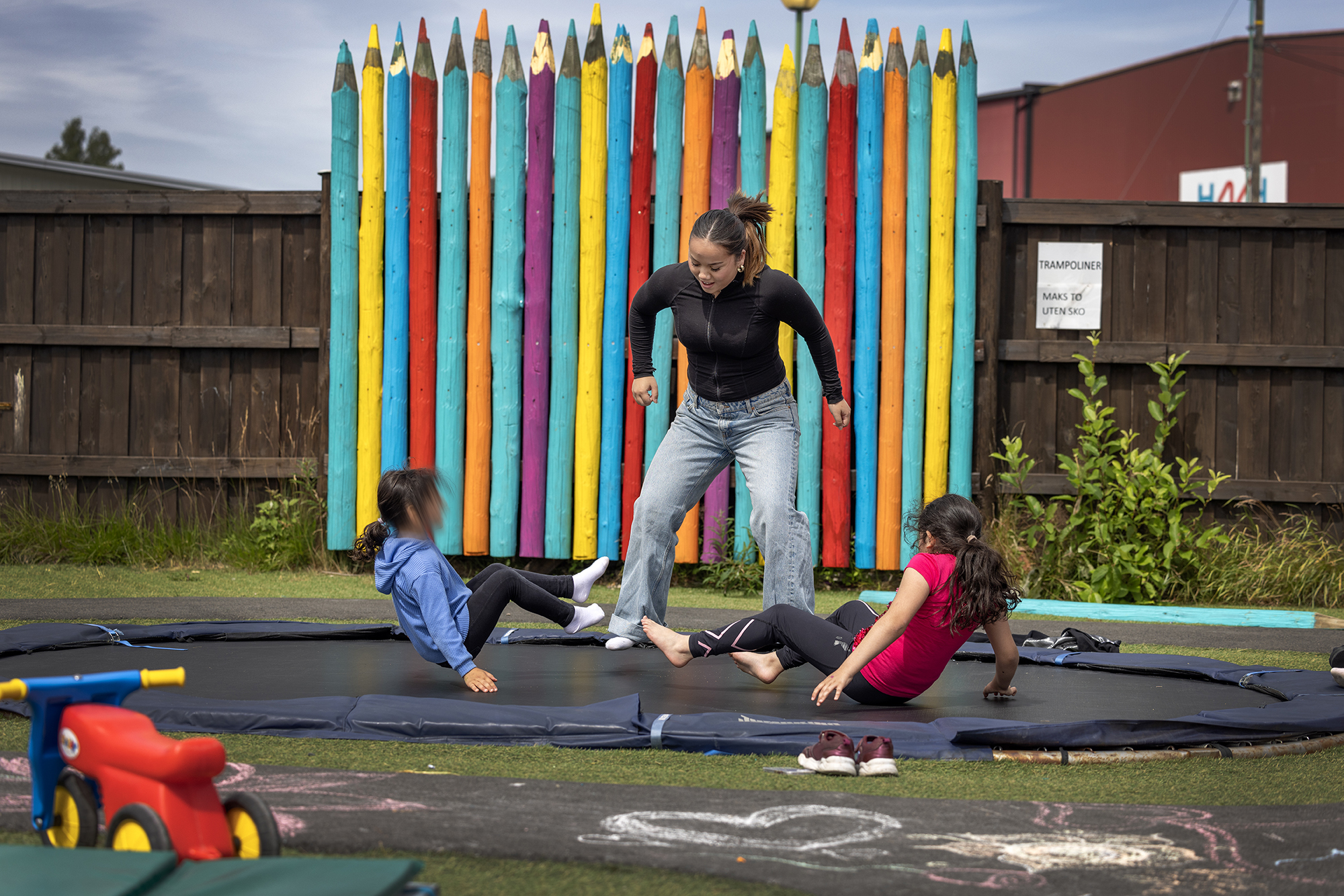 Foto: Tenåring som hopper på trampolina med to mindre barn. I bakgrunnen et gjerde som forestiller tegneblyanter.